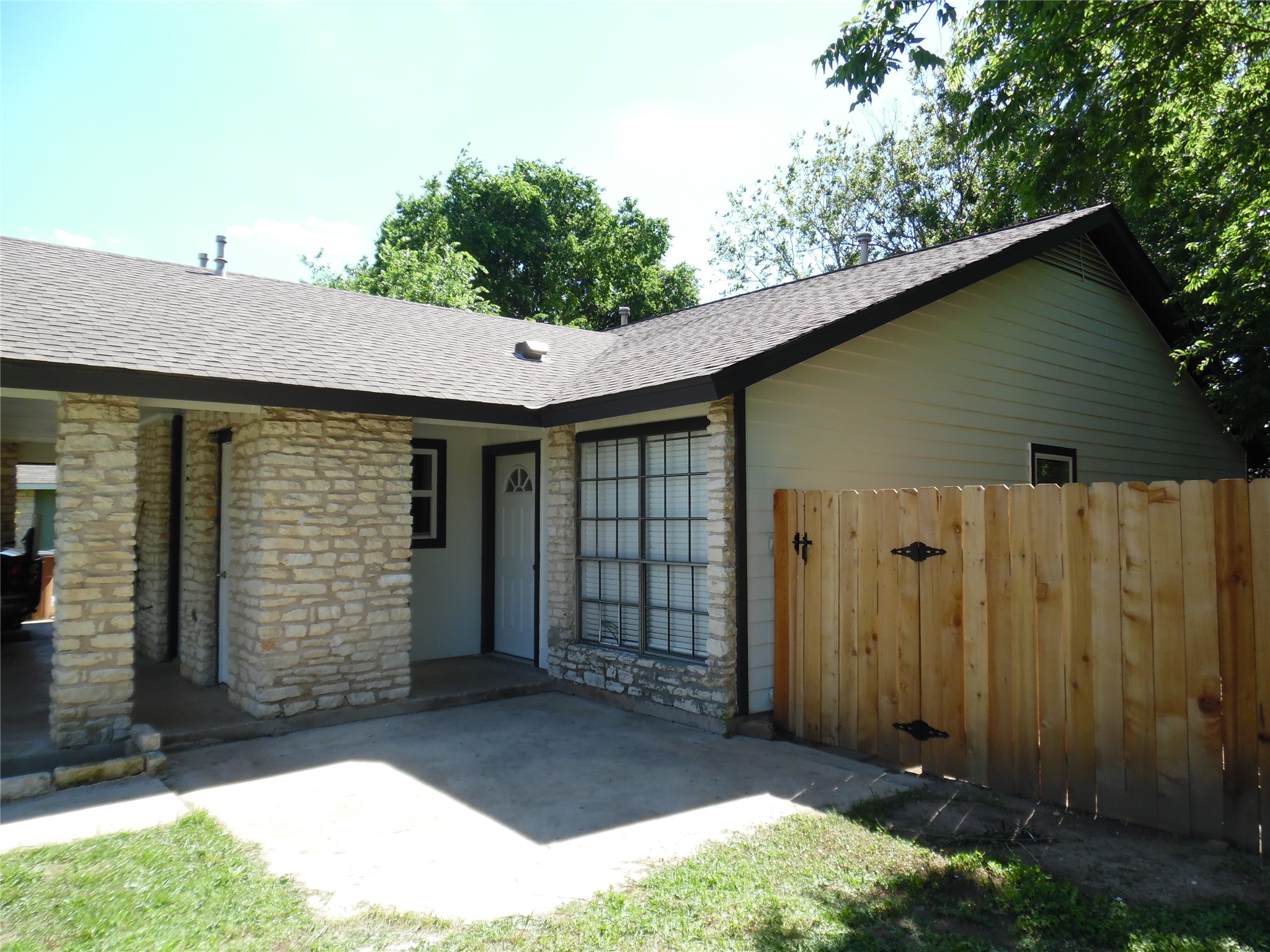 7400 Dixie Drive, Unit B Austin, TX 78744 - Photo 2 of 15 Stone facade home with a shingle roof, featuring an entry door, a window with horizontal blinds, and a wood privacy fence with decorative iron hardware