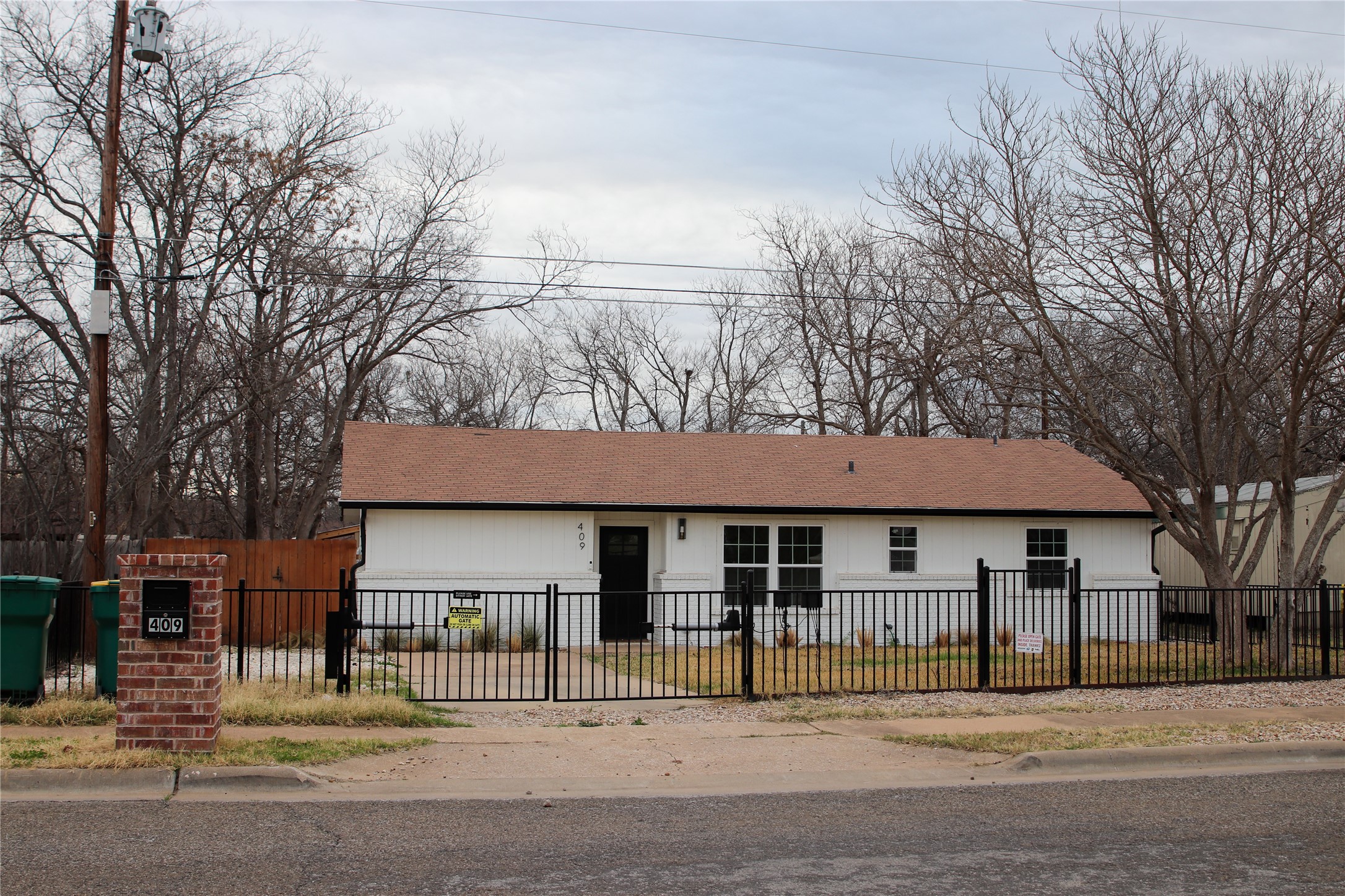 409 Ave R Marble Falls, TX 78654 - Photo 5 of 25 Photo taken in winter White siding residence with a brown shingle roof, featuring a black security fence and gate at the front