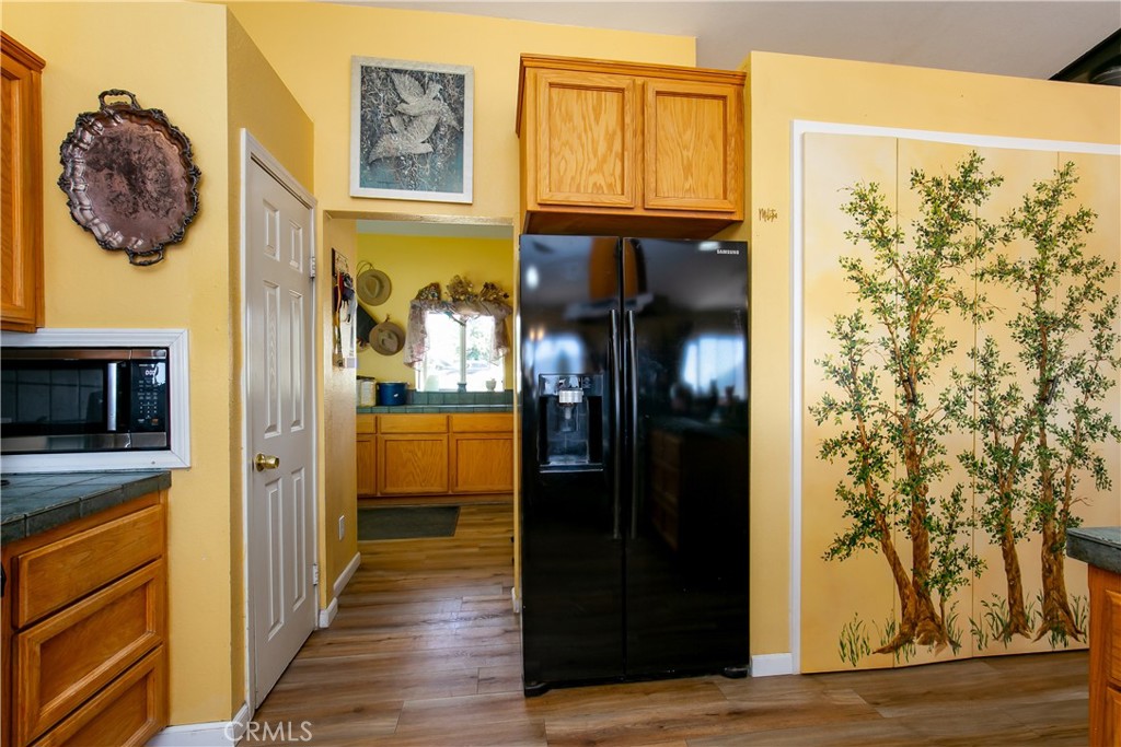 10960 Steinhoff Road Frazier Park, CA 93225 - Photo 28 of 46 A functional interior perspective showcasing the transition from the kitchen to the utility spaces. This shot highlights a large black refrigerator nestled against warm yellow cabinetry, leading down the hallway toward the laundry room entry, complemented by a unique painted tree mural on the walk-in pantry door.