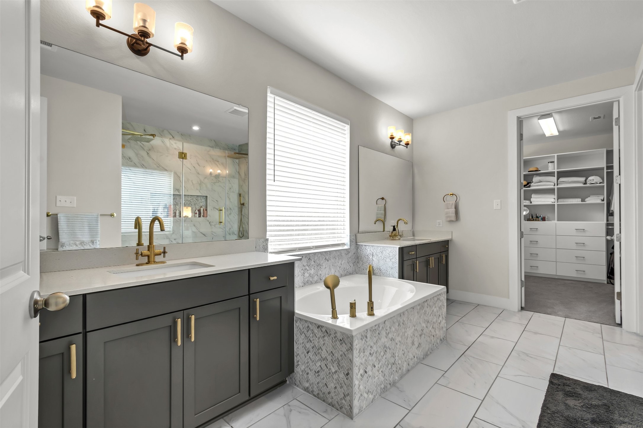 209 Azalea Spg Road Liberty Hill, TX 78642 - Photo 25 of 29 Spacious bathroom featuring dual vanities with quartz countertops, a tiled soaking tub with brass fixtures, a walk-in shower with glass enclosure, and an adjacent walk-in closet with built-in shelving