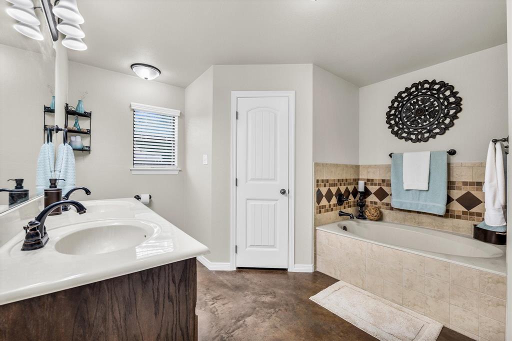 814 Springdale Circle Lorena, TX 76655 - Photo 29 of 37 Bathroom featuring a dual vanity with wood-finish cabinetry, a large soaking tub with tile surround, a window with blinds, and durable concrete-style flooring