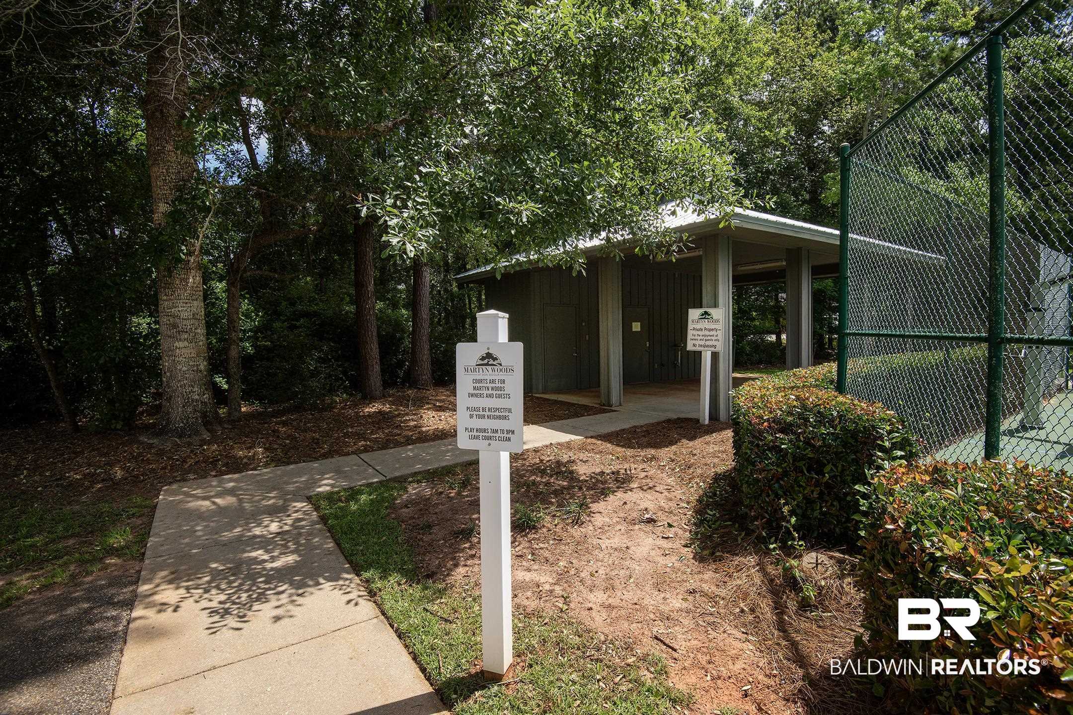18754 Treasure Oaks Road Gulf Shores, AL 36542 - Photo 45 of 53 Concrete pathway leading to a recreational building with a covered patio, surrounded by mature trees and manicured hedges, alongside a fenced tennis court