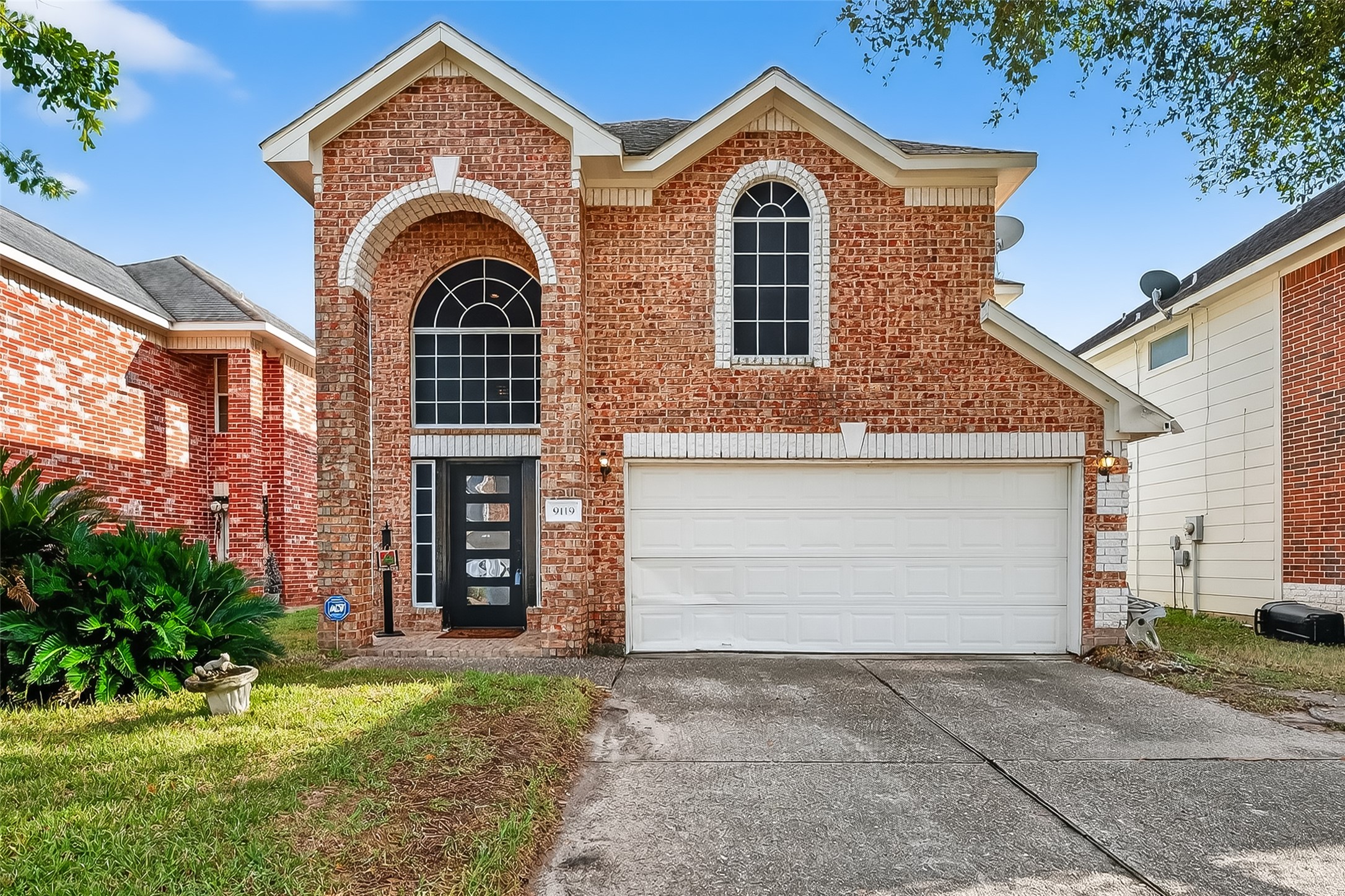 Charming two-story brick home with a classic arched window above the entrance. It features a two-car garage and a low-maintenance yard, set in a suburban neighborhood.