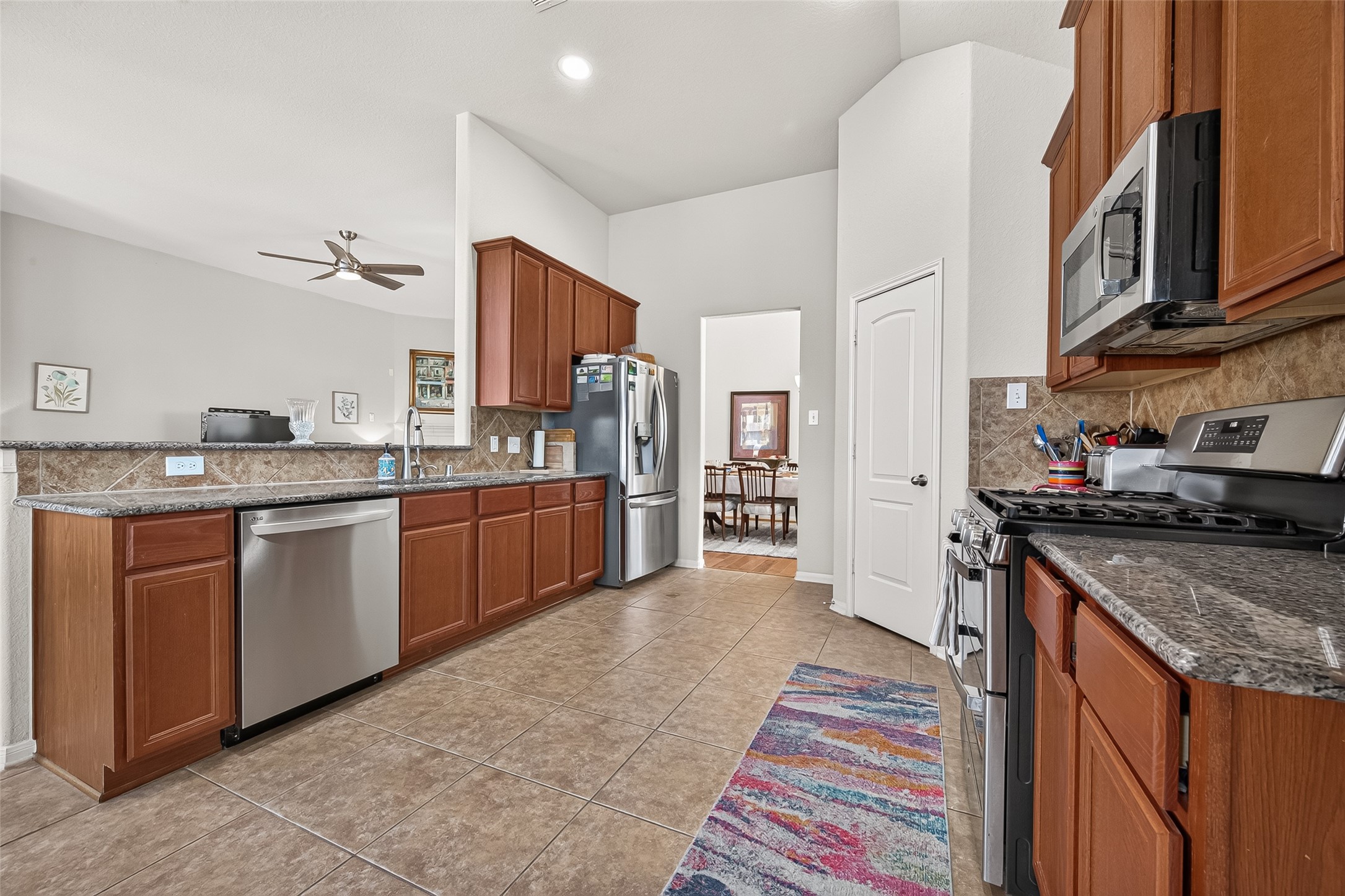 4910 Culver Lane Rosenberg, TX 77471 - Photo 26 of 50 This modern kitchen features stainless steel appliances, granite countertops, and ample wooden cabinetry. It has an open layout with a view into the dining area and living space, accented by a ceiling fan. The tile flooring and neutral walls create a welcoming atmosphere.