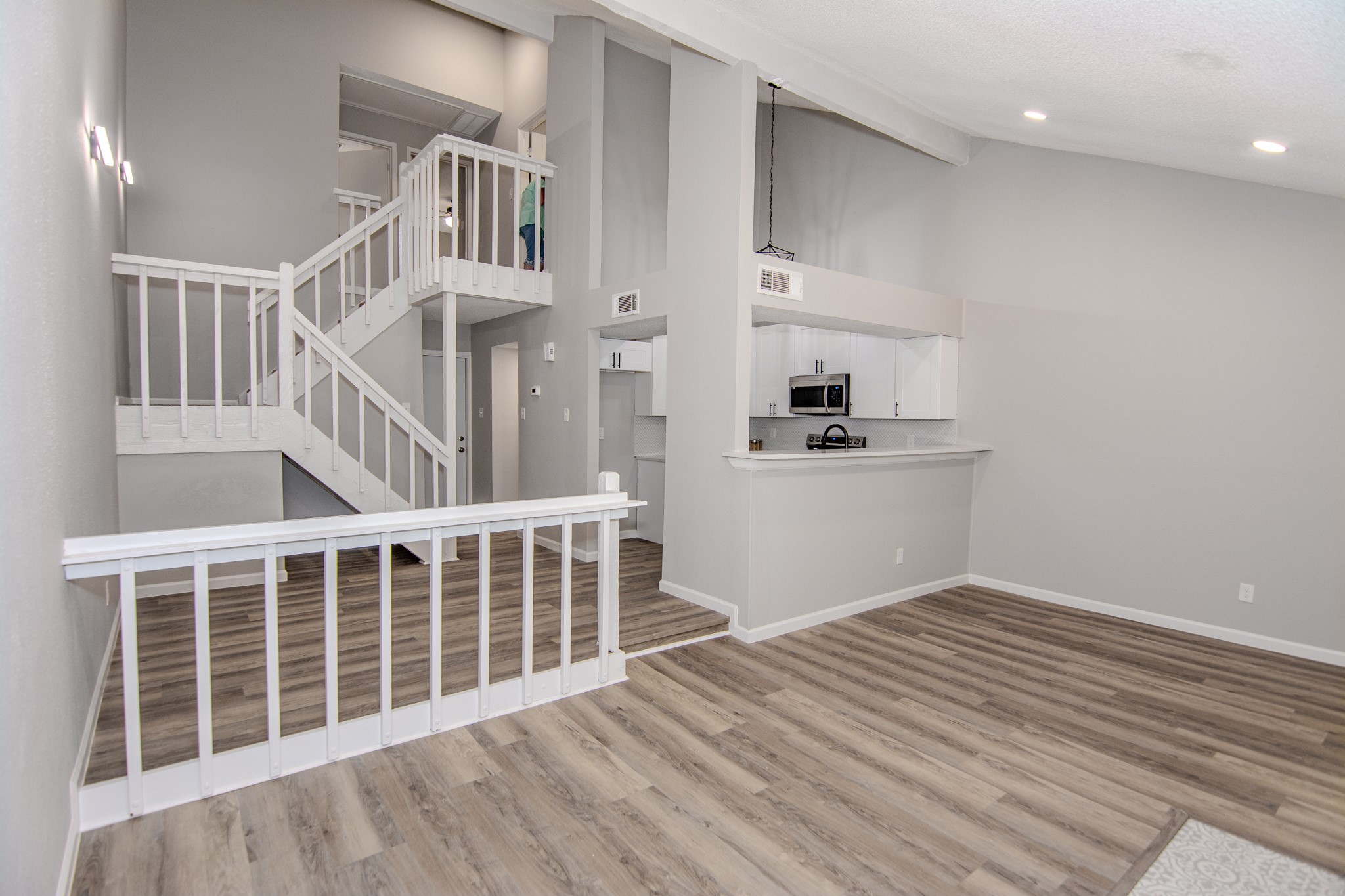 13009 Wirevine Lane Houston, TX 77072 - Photo 9 of 18 Modern, open-concept living area with high ceilings and a sleek staircase. It features light Vinyl plank wood flooring, neutral walls, and a small kitchen with a breakfast bar and stainless steel appliances, offering a spacious and contemporary feel.