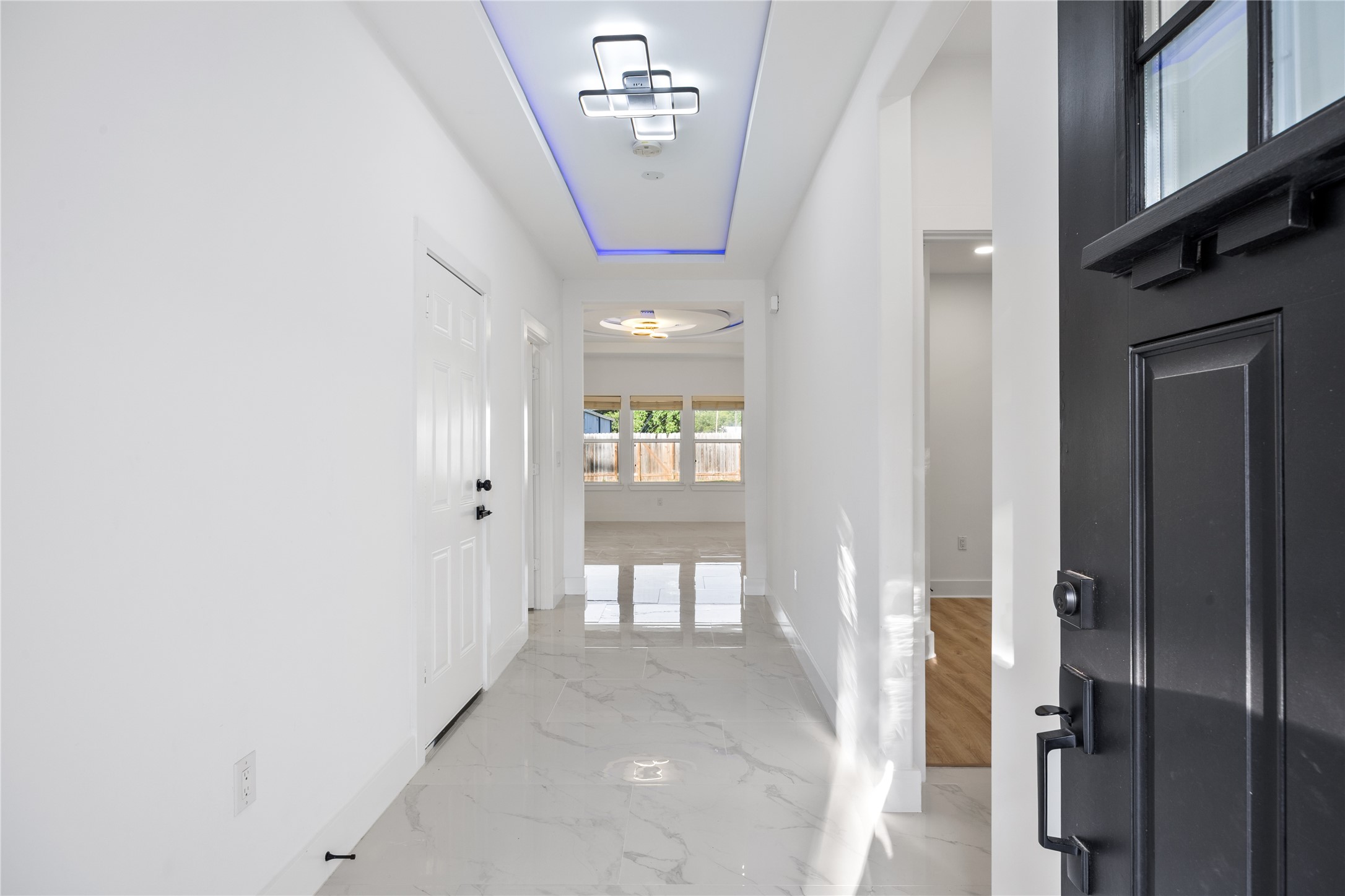 1121 Austin Street Rosenberg, TX 77471 - Photo 5 of 44 This photo showcases a modern, bright entryway with glossy quartz floors and white walls. The hallway features contemporary lighting with a blue accent and leads to a spacious living area with large windows overlooking a backyard.