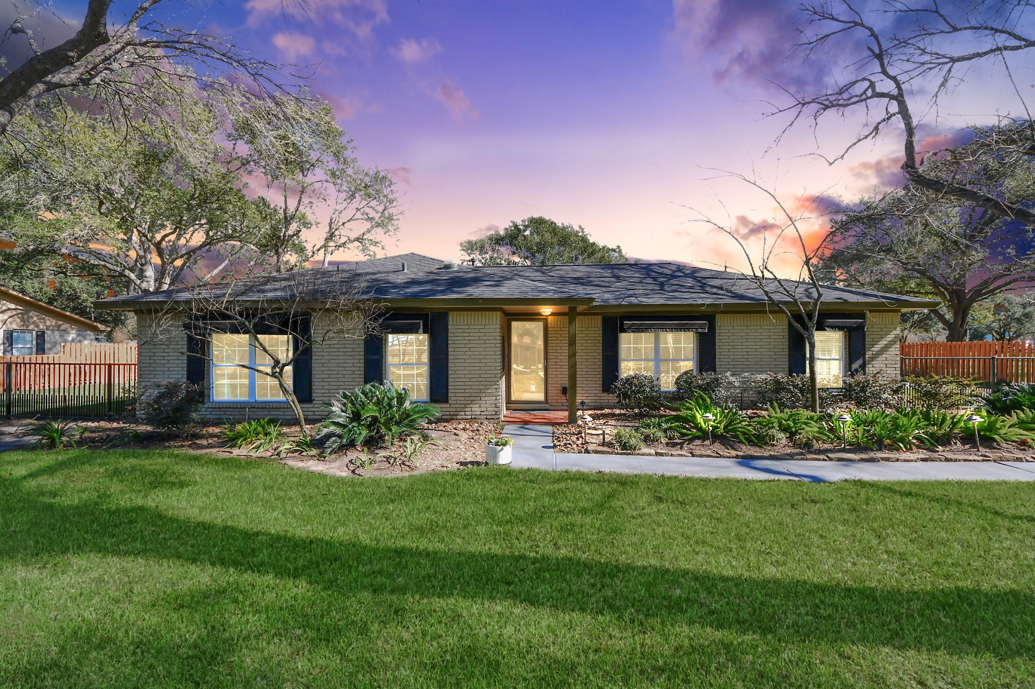 This charming single-story home features a welcoming front yard with lush greenery, mature trees, and a well-maintained lawn. The house has a traditional design with a brick exterior and large windows, providing a warm and inviting appearance. A spacious driveway and a cozy front porch complete the picturesque setting.