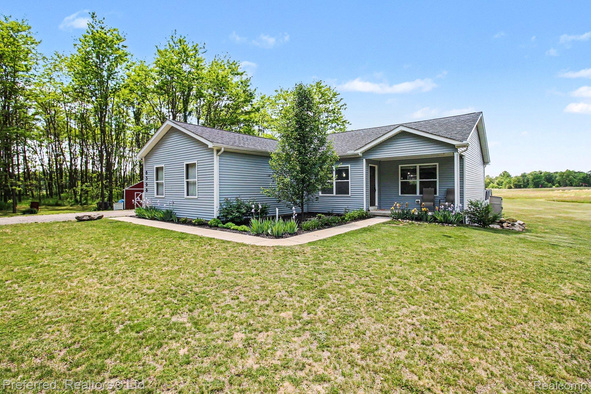 6366 Dutcher Road Howell, MI 48843 - Photo 1 of 33 Light blue siding exterior with a dark shingle roof, featuring a covered front porch, a concrete walkway, and landscaped garden beds
