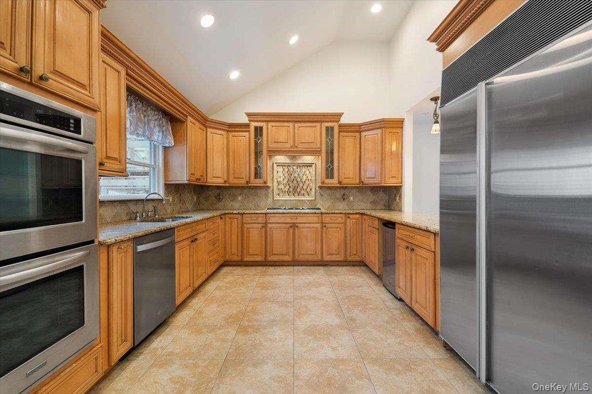 33 Fleets Cove Road Huntington, NY 11743 - Photo 3 of 42 Main Kitchen featuring appliances with stainless steel finishes, light stone countertops, high vaulted ceiling, decorative backsplash, and brown cabinetry