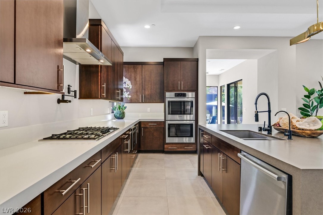 53 Garibaldi Way Henderson, NV 89011 - Photo 21 of 80 Kitchen with appliances with stainless steel finishes, wall chimney exhaust hood, dark brown cabinets, a kitchen island with sink, and light tile patterned floors