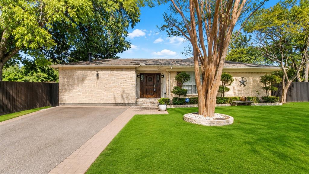 10505 Ferguson Road Dallas, TX 75228 - Photo 1 of 14 Stone facade home featuring a prominent front lawn, mature trees, a paved driveway with brick paver edging, and a dark wooden privacy fence