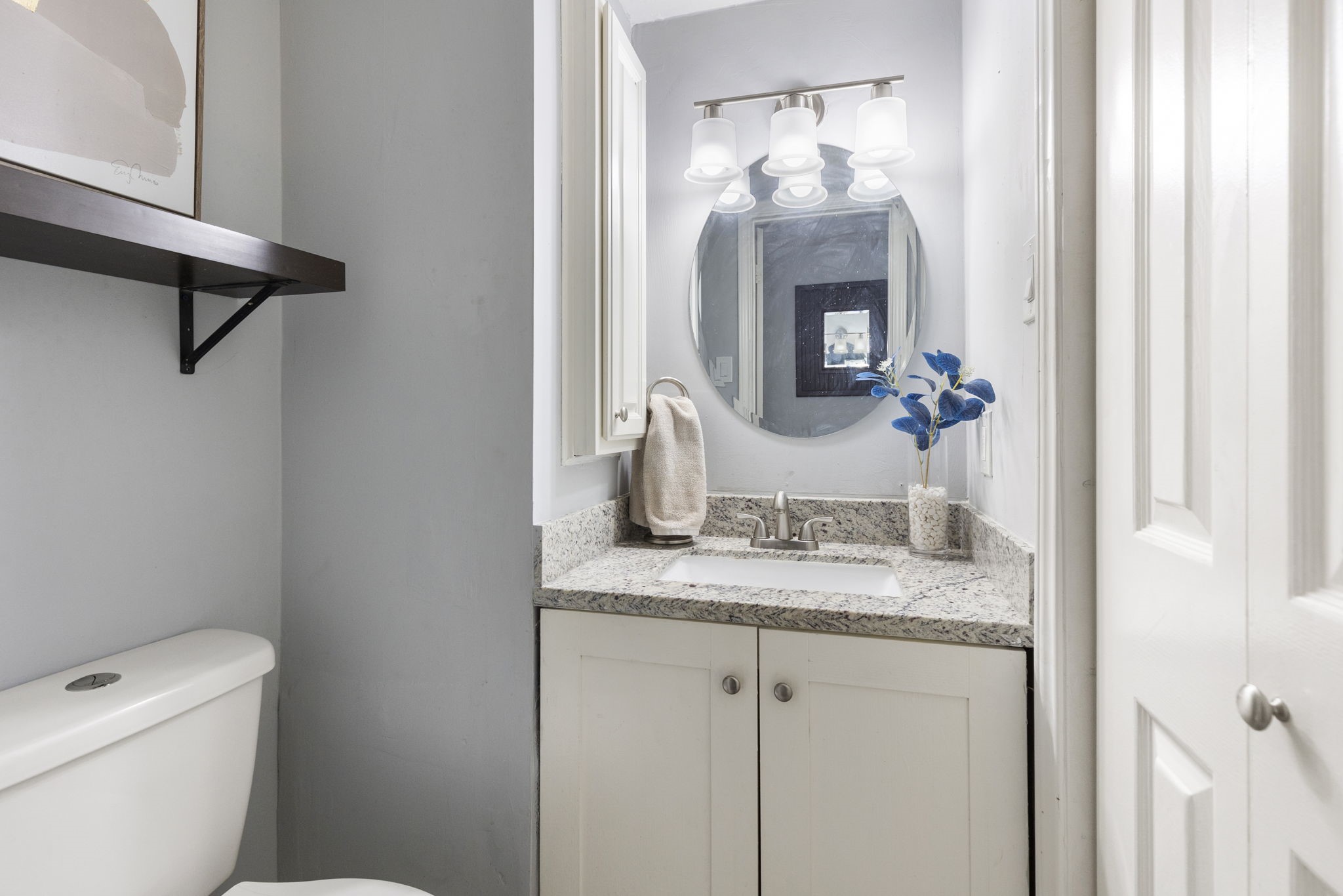 2110 Paso Rello Drive Houston, TX 77077 - Photo 19 of 41 This is a sleek, modern bathroom featuring a granite countertop, a round mirror, and stylish lighting. It includes a compact vanity, a toilet, and a decorative shelf, creating a clean and inviting space.