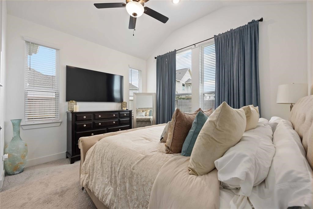 10014 Nectar Path Iowa Colony, TX 77583 - Photo 18 of 26 This bedroom features a cozy, carpeted space with a ceiling fan, natural light from large windows with blue curtains, and a stylish setup including a TV, dresser, and plush bedding.