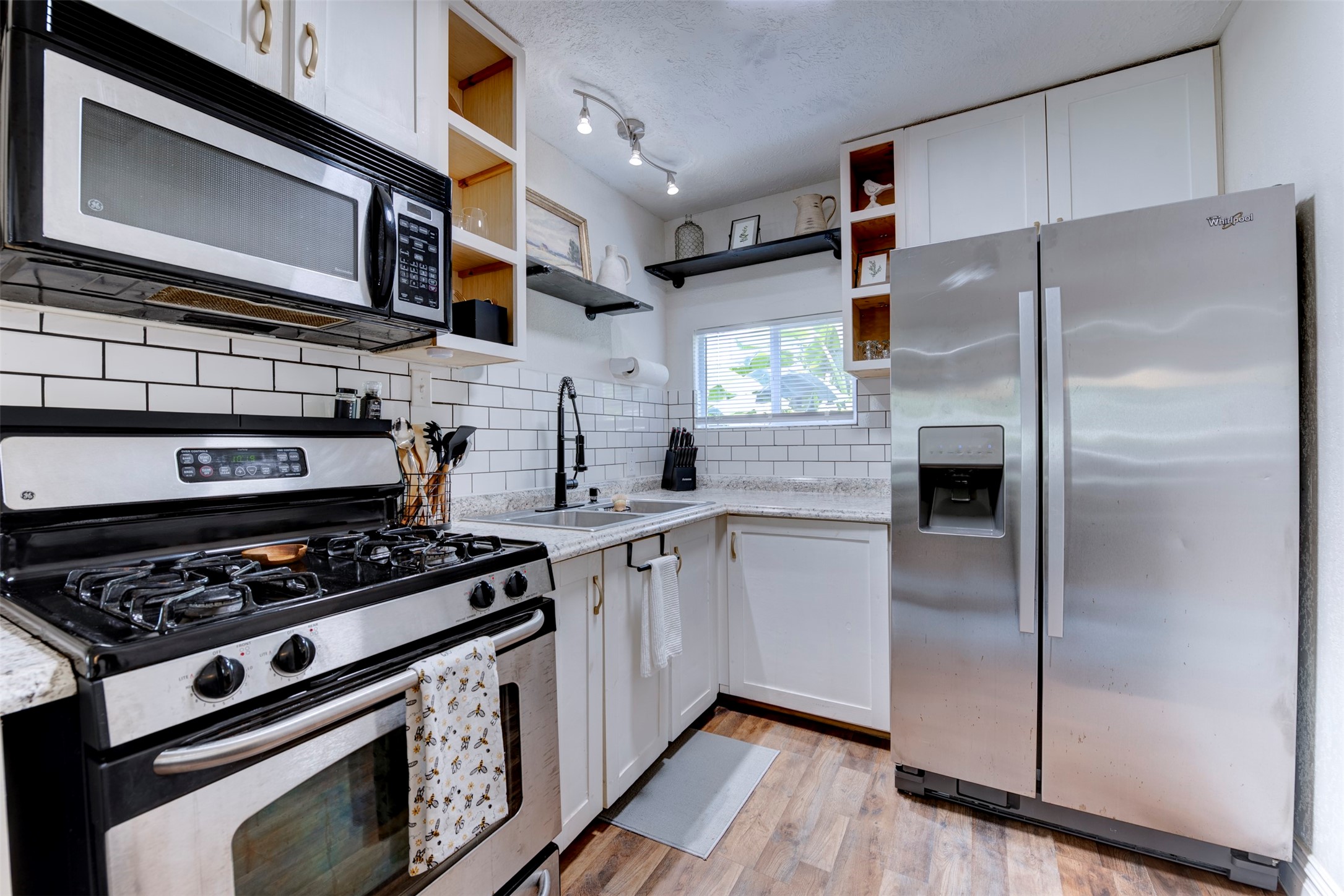 1121 Panama Street Houston, TX 77009 - Photo 11 of 35 Full kitchen view featuring GE five-burner gas range with stainless finish, over-the-range stainless microwave, Whirlpool side-by-side refrigerator with ice and water dispenser, white shaker cabinetry with brushed gold hardware, granite countertops, white subway tile backsplash with dark grout, open pine cubby shelving, black pipe bracket floating shelf, track lighting, and operable window.