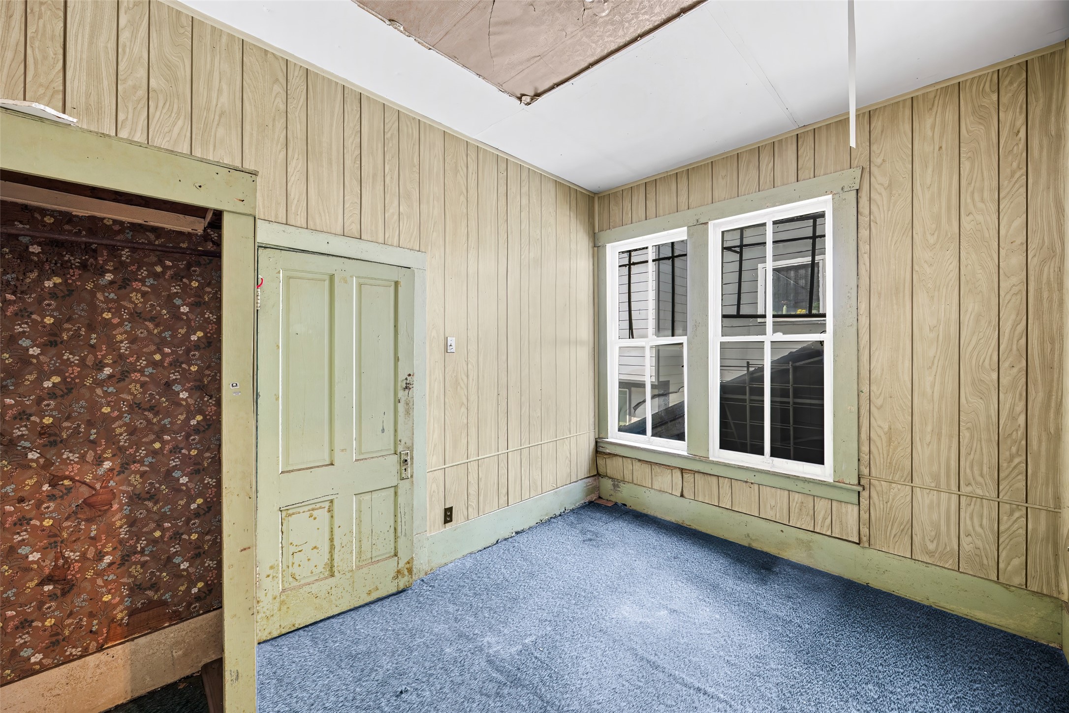 1214 Edwards Street Houston, TX 77007 - Photo 14 of 34 This room features wood paneling, blue carpet, and a vintage green door with a floral-patterned closet. Natural light filters through two large windows, adding potential for a cozy, refreshed space.