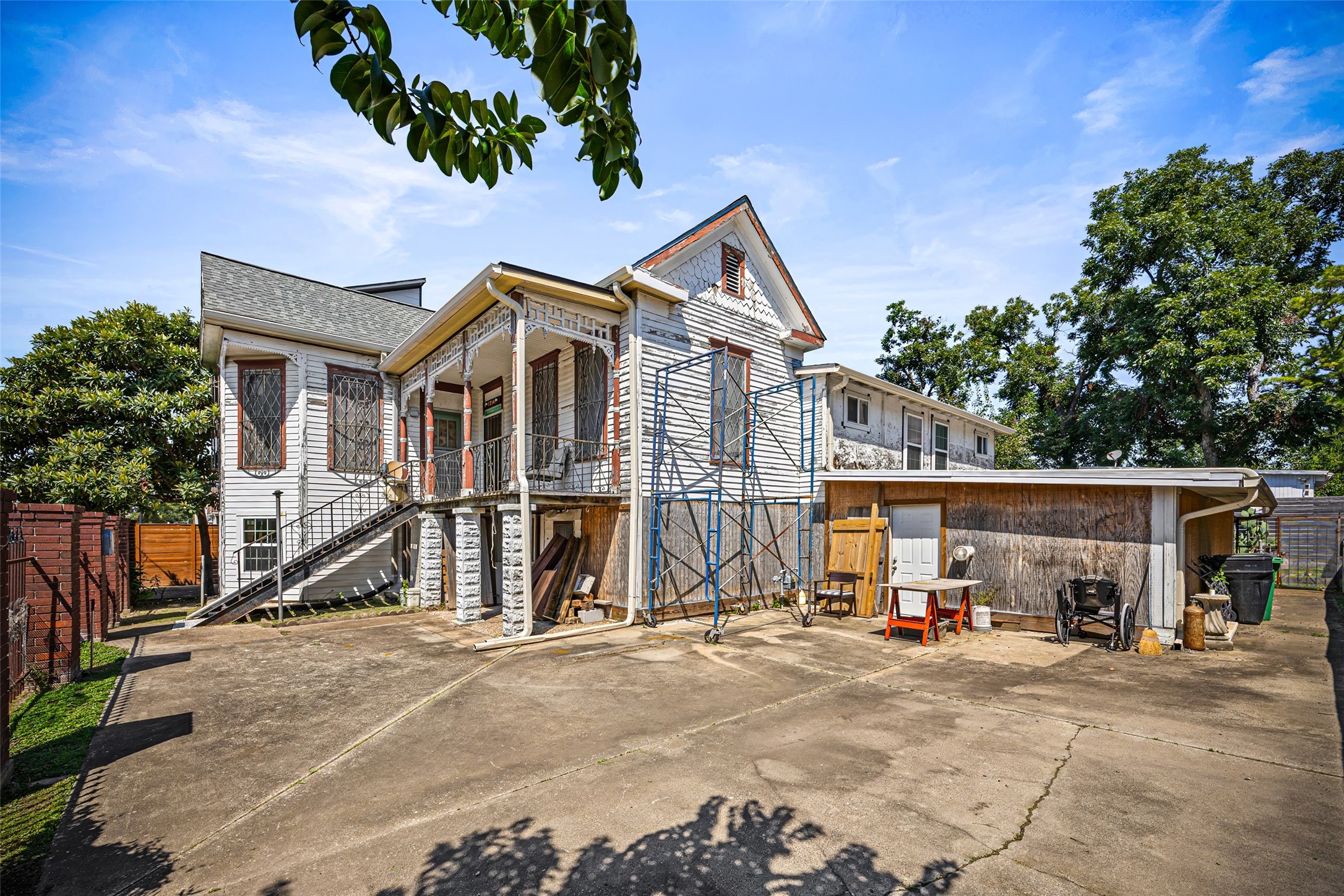 1214 Edwards Street Houston, TX 77007 - Photo 6 of 34 This photo features a two-story house with a rustic exterior, surrounded by mature trees. It has a spacious concrete yard area, a covered porch with intricate railing, and a side stairway. The property appears to have potential for customization or renovation.