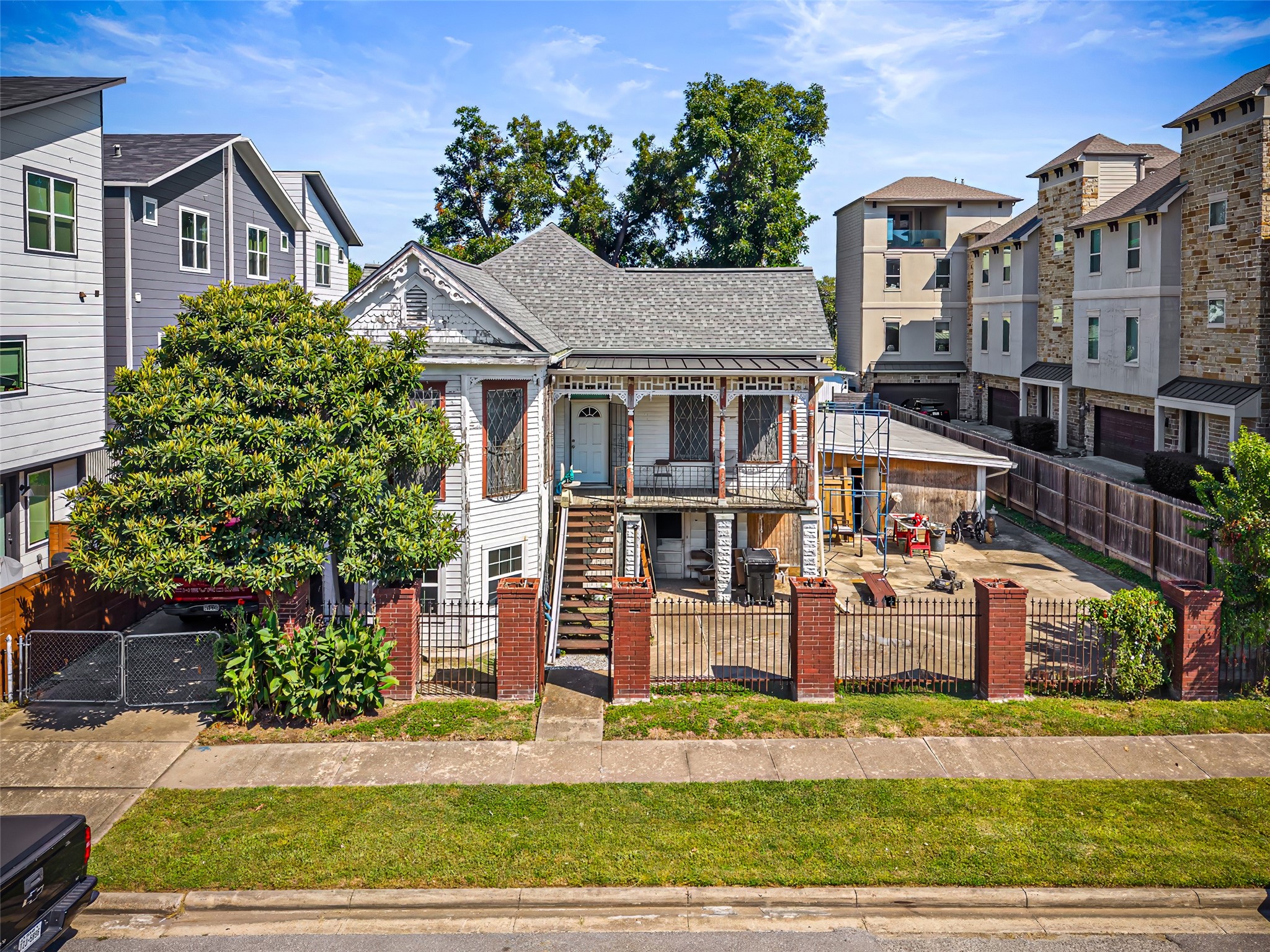 1214 Edwards Street Houston, TX 77007 - Photo 7 of 34 This photo features a charming older home with intricate architectural details, set behind a brick and iron fence. The house has a spacious front porch, a mature tree, and a driveway leading to a garage. It's surrounded by a mix of modern and traditional neighboring homes, offering a blend of historical charm and contemporary living.