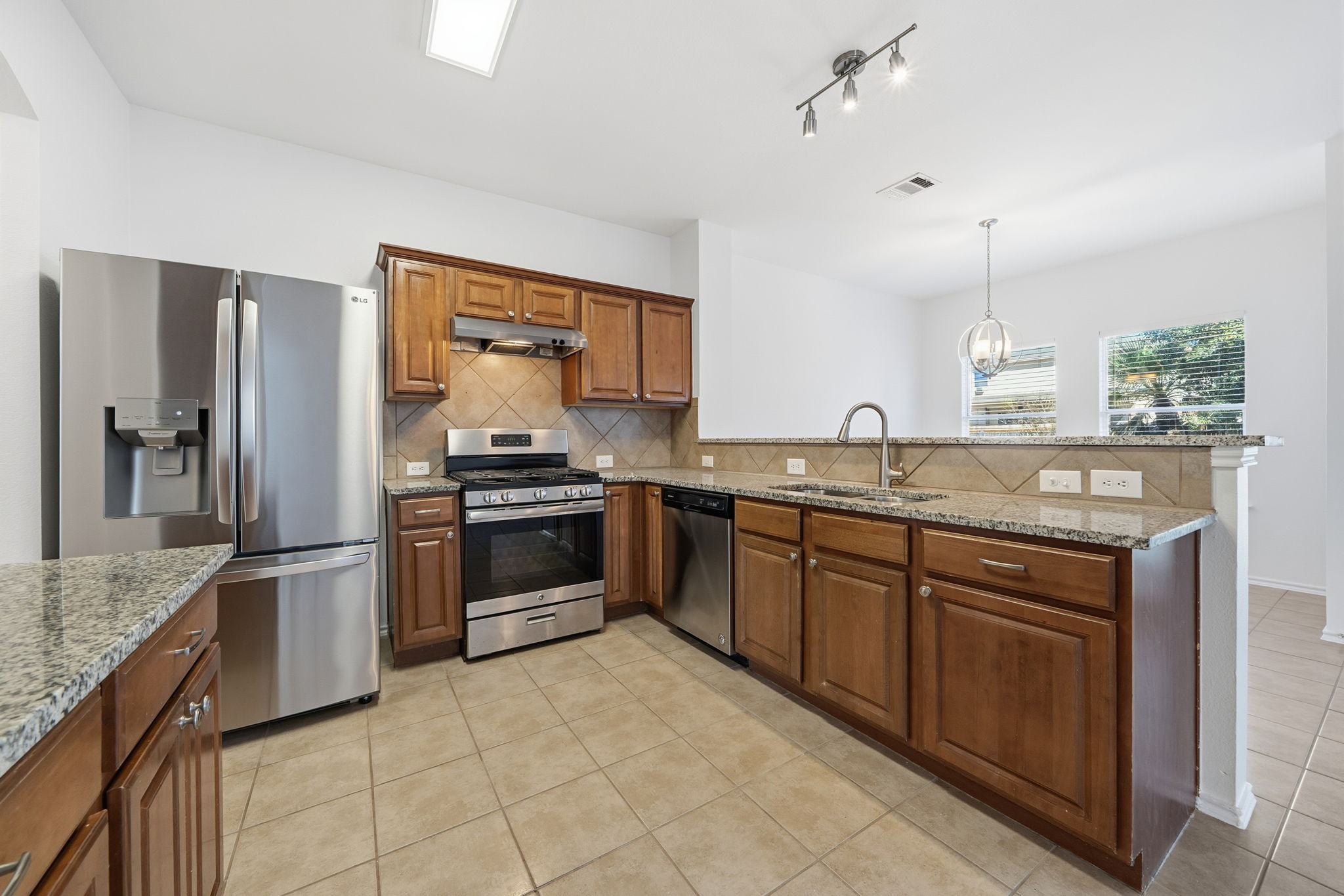 5712 Zachary Scott Street Austin, TX 78747 - Photo 6 of 28 Kitchen with appliances with stainless steel finishes, brown cabinetry, light tile patterned flooring, a peninsula, and light stone counters
