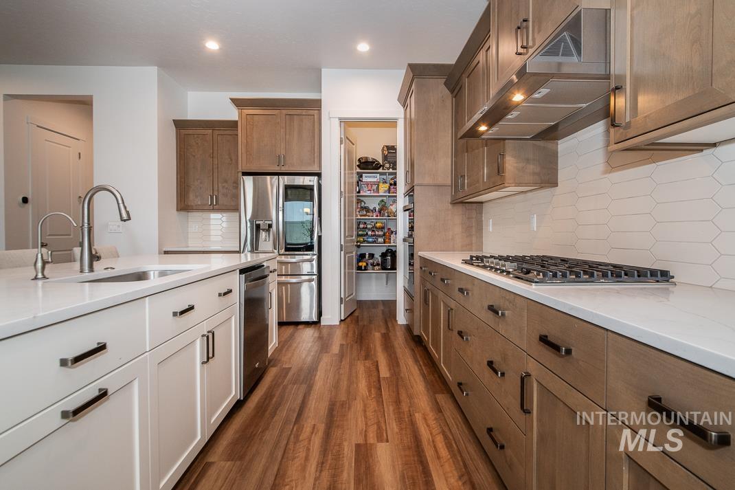 5610 Sparky Avenue Caldwell, ID 83607 - Photo 6 of 14 Kitchen featuring under cabinet range hood, brown cabinetry, tasteful backsplash, dark wood-style flooring, and appliances with stainless steel finishes