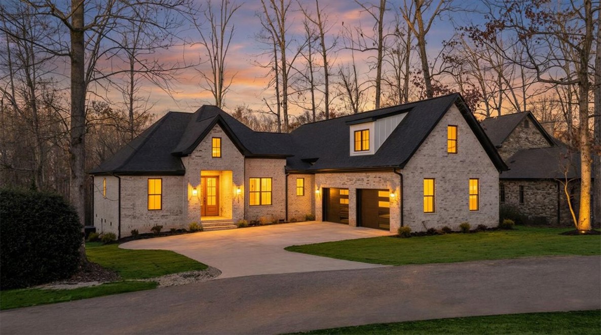 This modern home features a brick exterior, an attached garage, and a long driveway surrounded by natural landscaping.