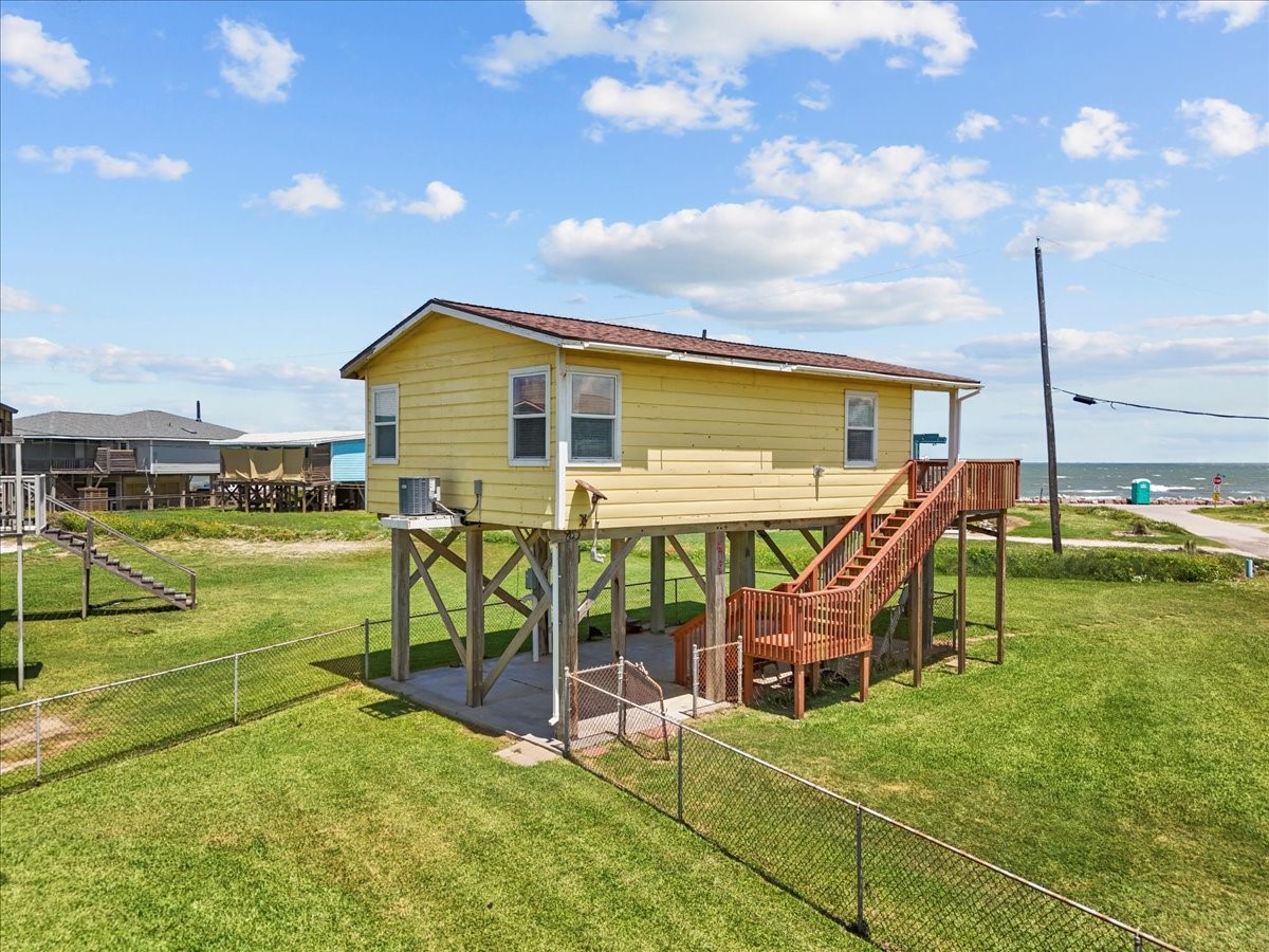 819 Seashell Drive Surfside Beach, TX 77541 - Photo 31 of 36 Elevated yellow beach house with ocean views, surrounded by a grassy yard and fenced area. Features a wooden staircase leading to the main entrance.