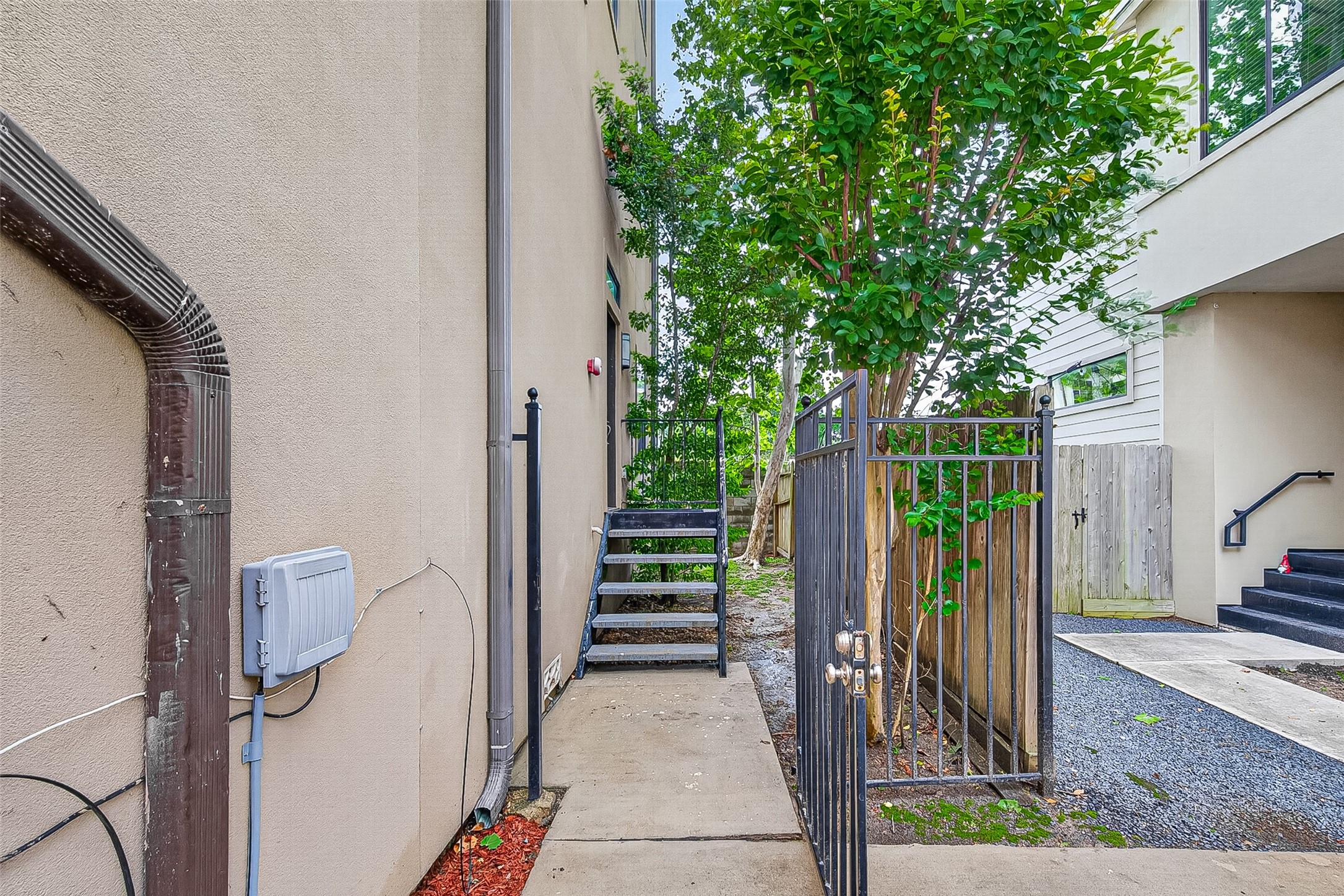 2611 Riverside Drive, Unit C Houston, TX 77004 - Photo 43 of 50 This photo shows a side pathway of a home with a concrete walkway, metal gate, and stairs leading up to a side entrance. There's a small garden area with a tree and some gravel landscaping. The exterior is modern with beige walls.