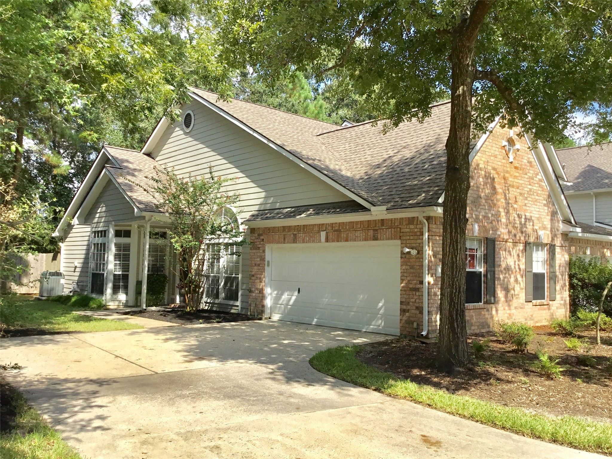 Charming single-story home with a brick and siding exterior, featuring a two-car garage. The property is surrounded by mature trees, offering a shaded front yard and a welcoming driveway.