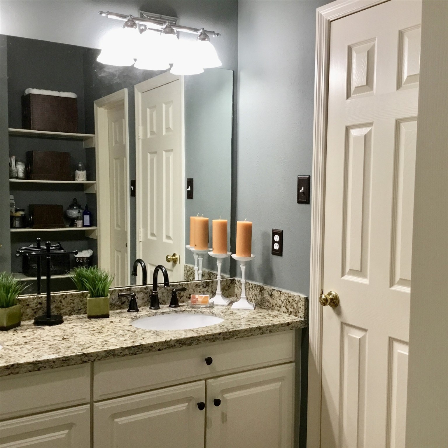 3 North Spiral Vine Circle Spring, TX 77381 - Photo 22 of 41 This bathroom features a granite countertop with a single sink, white cabinetry, and modern fixtures. It includes a large mirror, soft lighting, and shelves for storage, creating a cozy and functional space.