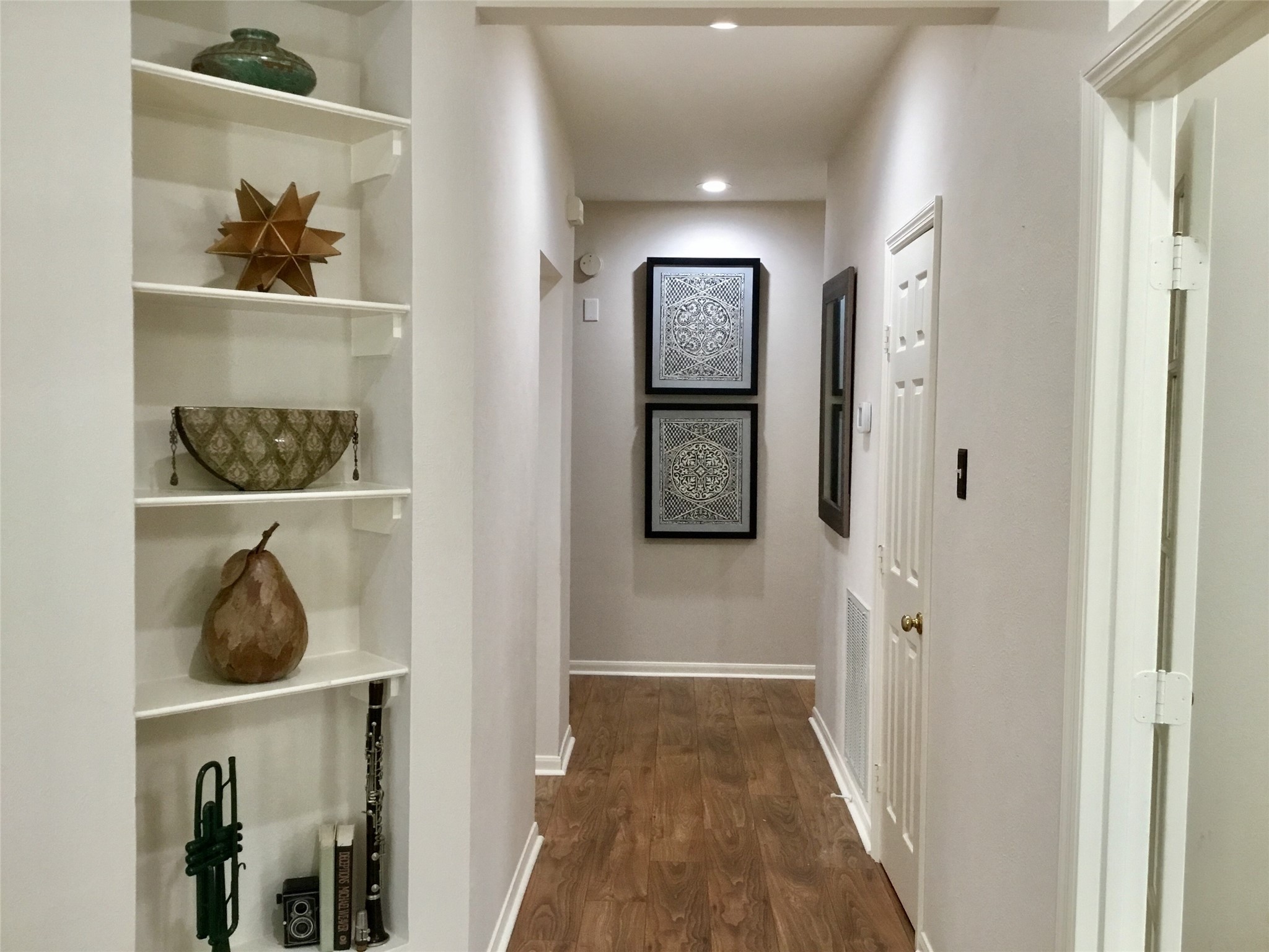 3 North Spiral Vine Circle Spring, TX 77381 - Photo 3 of 41 This photo showcases a hallway with warm wood flooring and soft lighting. The left side features built-in shelves with decorative items, while the right wall displays framed artwork. The space feels inviting and well-lit.
