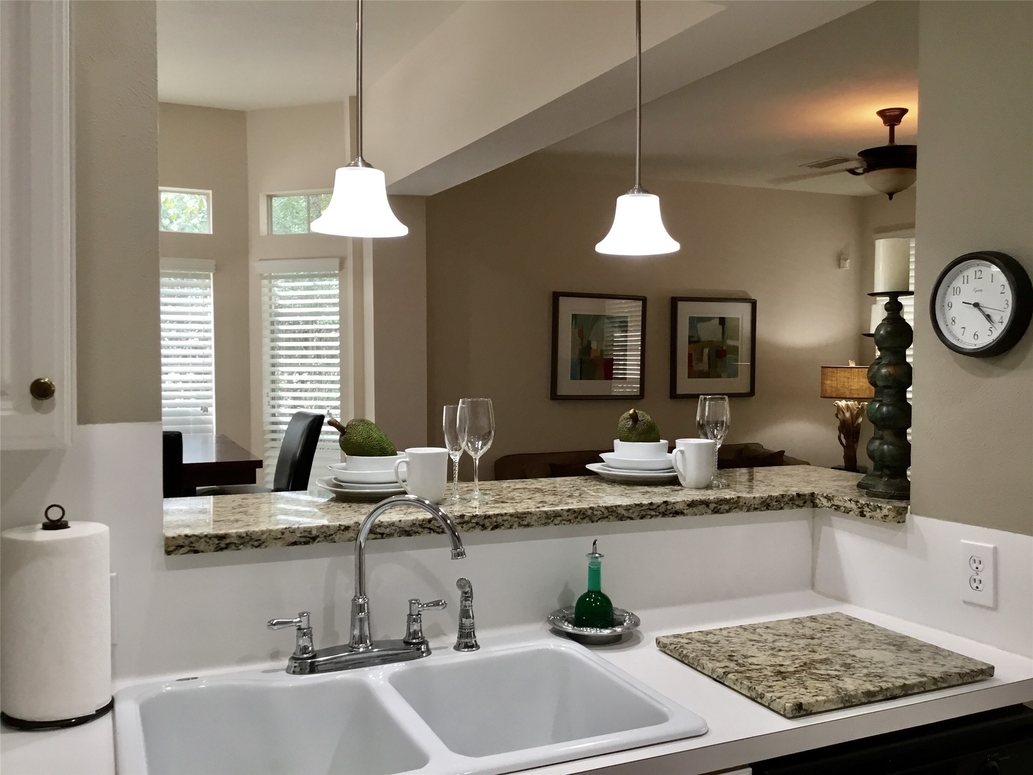 3 North Spiral Vine Circle Spring, TX 77381 - Photo 10 of 41 This photo showcases a modern kitchen with granite countertops and a dual sink. It features pendant lighting and an open design leading to a cozy living area with decor and art on the walls. Natural light enters through windows with white shutters.