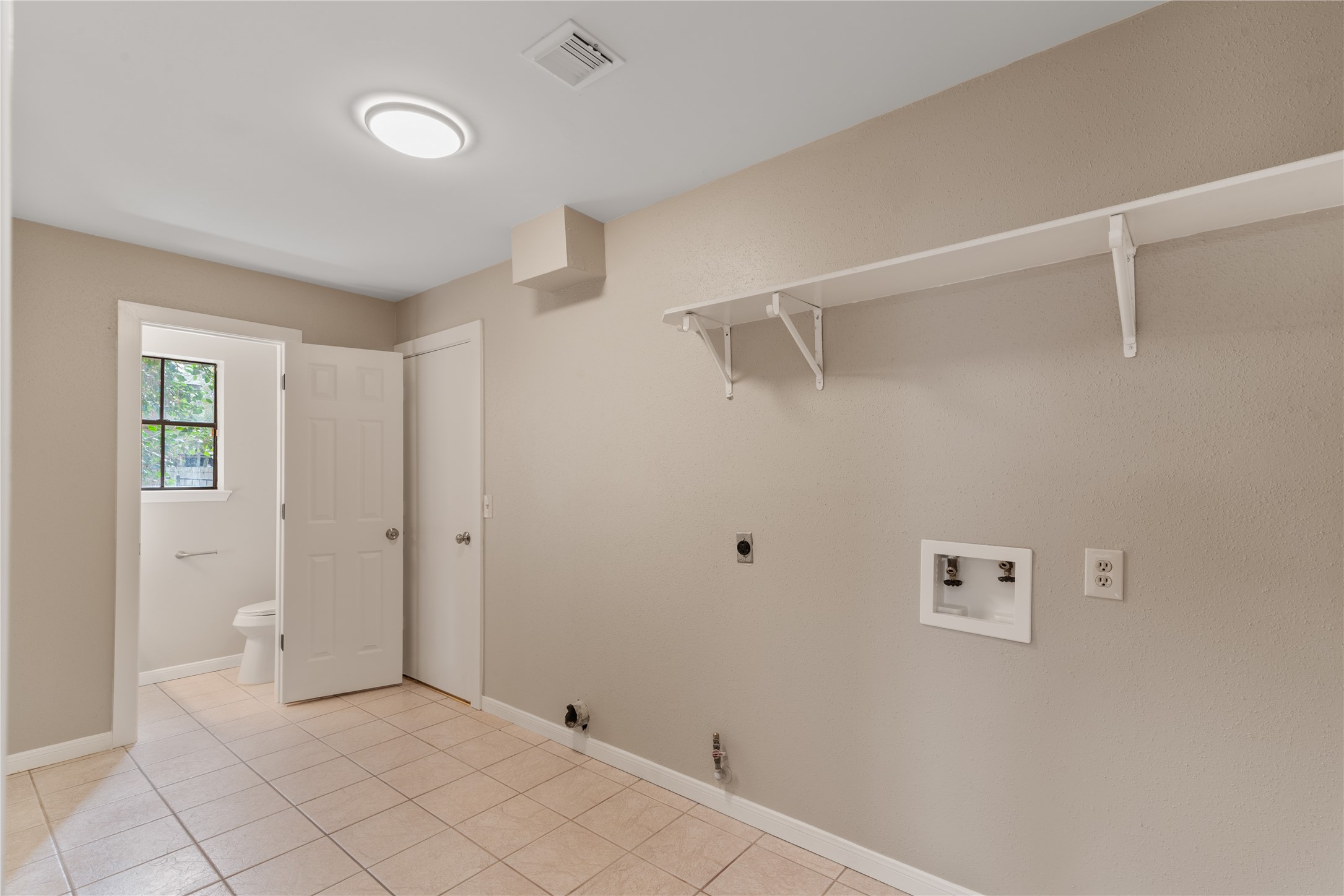 10 White Fawn Drive Spring, TX 77381 - Photo 14 of 25 This photo shows a utility room with neutral walls and tiled flooring. It includes hookups for a washer and dryer, a shelf for storage, and an adjacent half bath with a window.
