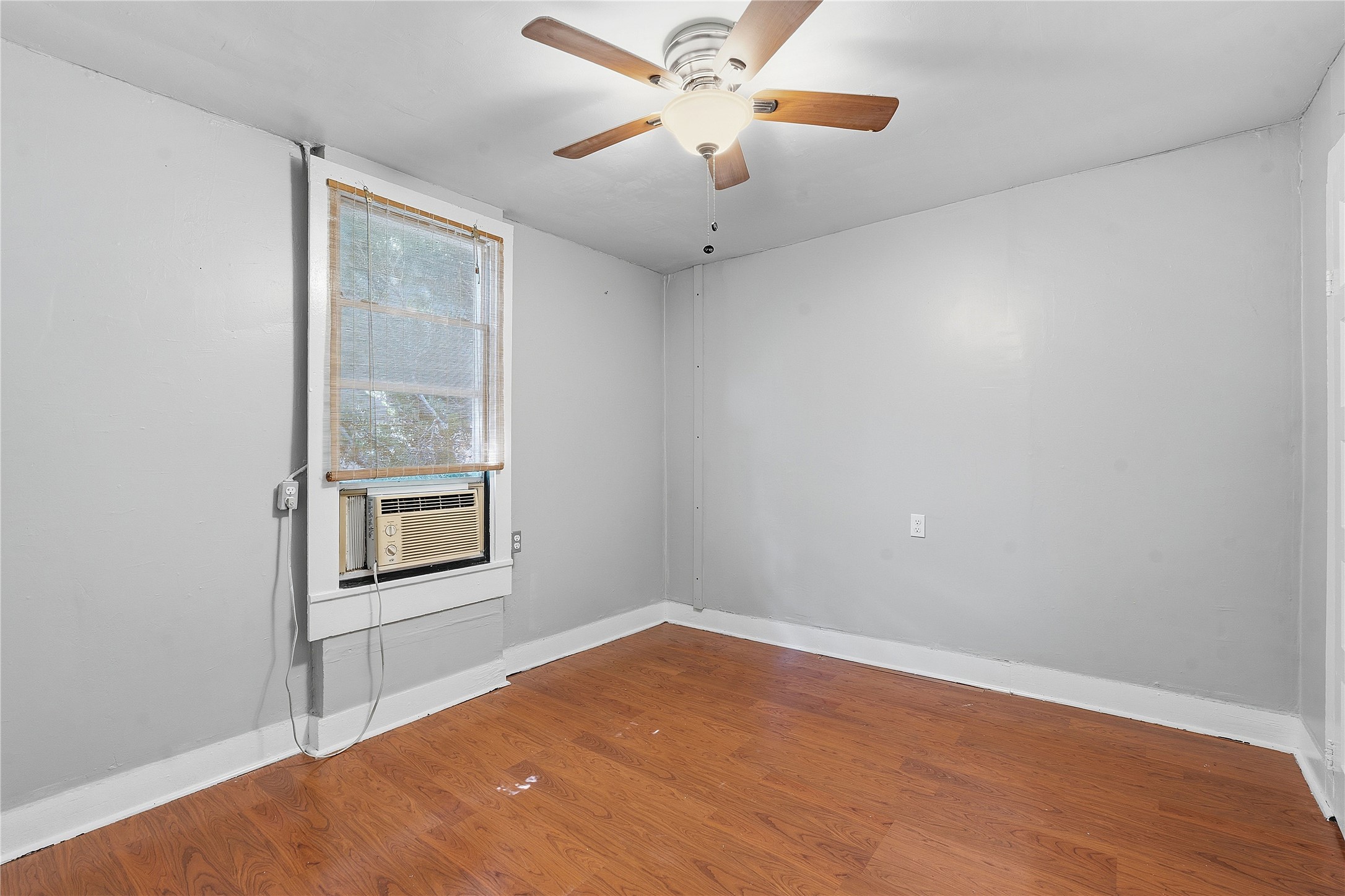 2207 Santa Rita Street Austin, TX 78702 - Photo 10 of 15 Room featuring wood-finish flooring, light gray walls, white trim, a window with a window-mounted AC unit, and a ceiling fan with integrated lighting