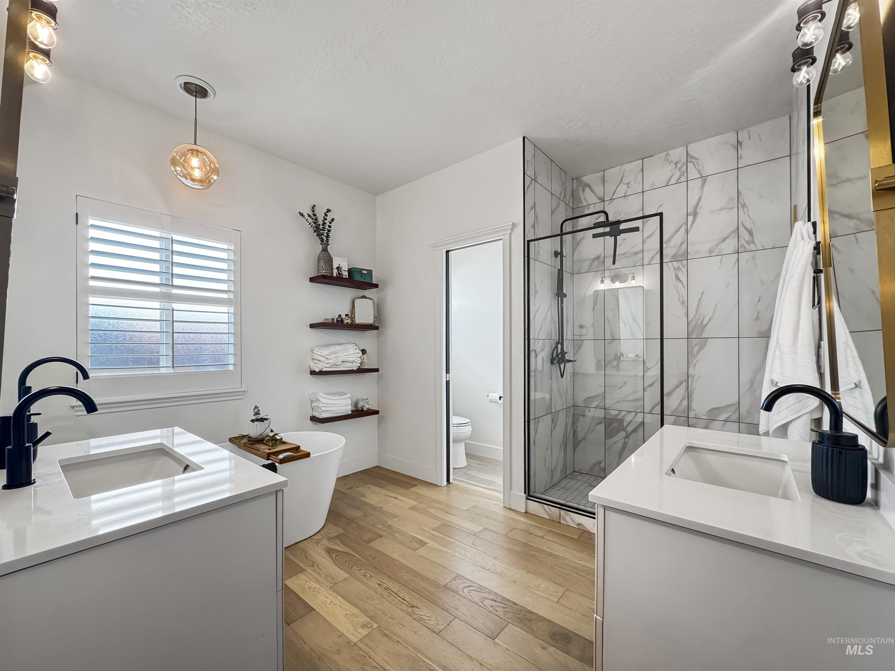3917 North Waterloo Way Boise, ID 83713 - Photo 13 of 24 Dual vanities with integrated sinks and matte black fixtures, a freestanding soaking tub, wood-finish flooring, a glass-enclosed shower with tiled walls, and a window with plantation shutters