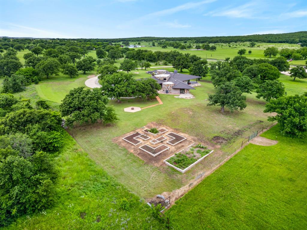 1080 North Greenbrier Road Nocona, TX 76255 - Photo 34 of 40 Expansive property featuring a contemporary residence with a dark roof and light-toned siding, mature trees, a curved driveway, and a fenced garden area with raised beds