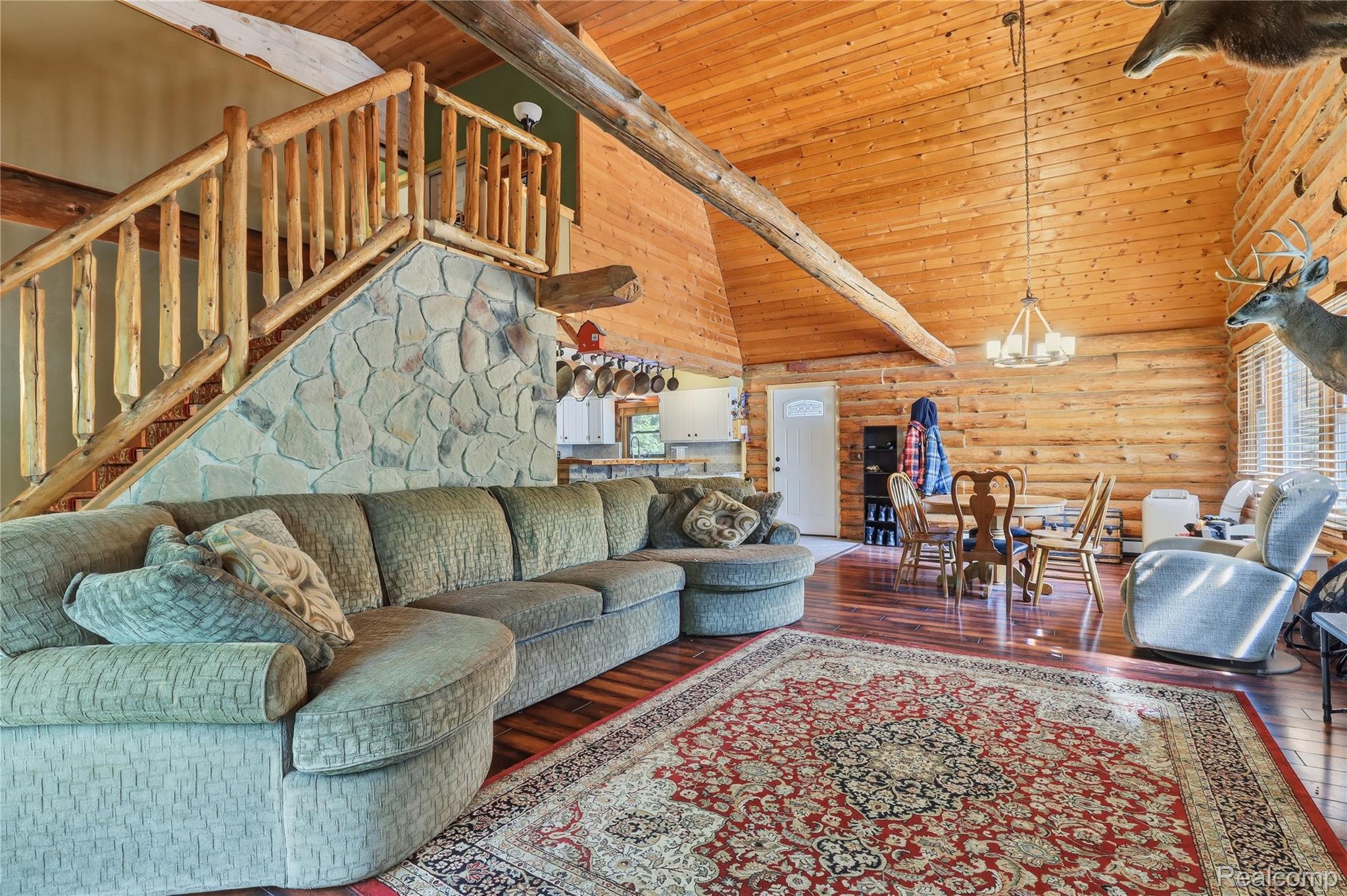 Open-concept living area featuring a vaulted wood-plank ceiling with exposed beams, wood-finish flooring, a natural stone staircase with a wood balustrade, and a dining area with an overhead chandelier