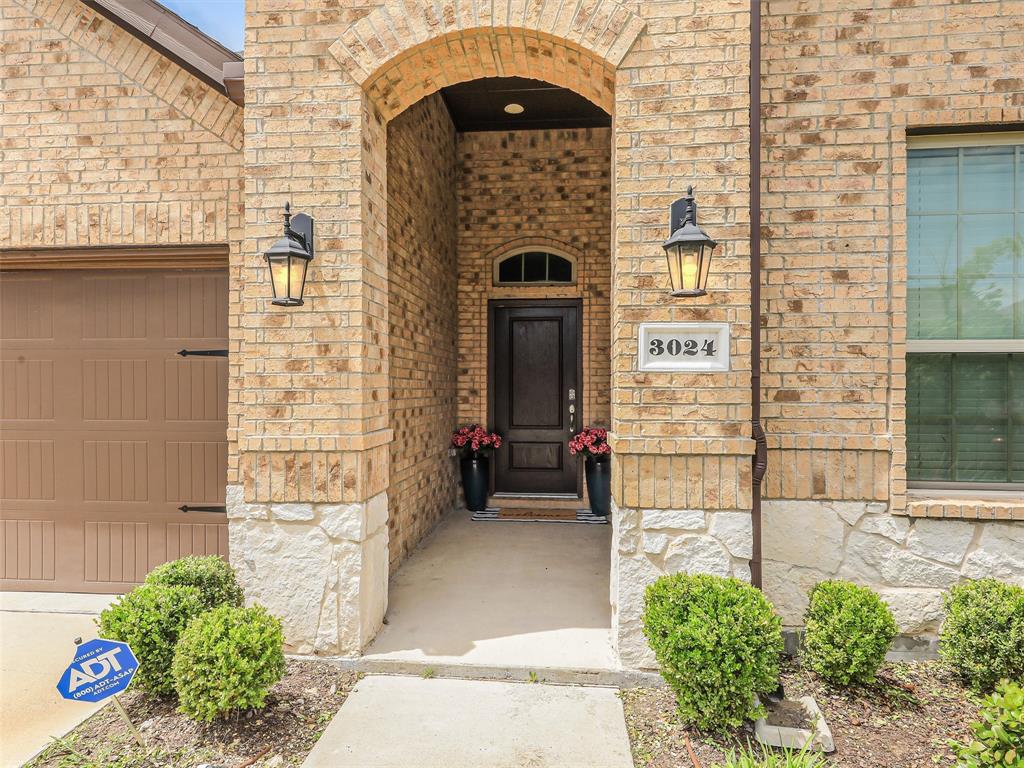 3024 Meredith Lane Forney, TX 75126 - Photo 2 of 29 Brick and stone exterior featuring an arched entryway with a dark wood-finish door, twin exterior lanterns, and a paneled garage door