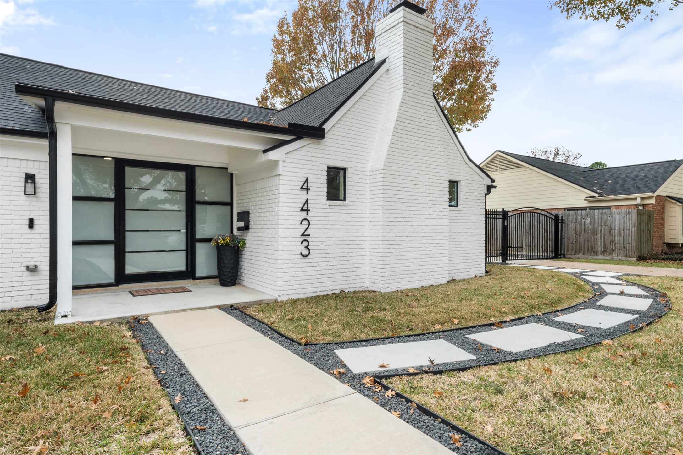 Welcome to 4423 Lymbar Drive! Exterior view showcasing white brick façade, large aluminum pivot glass door with surrounding windows, and stone landscaping accenting the walkway to the front entry and connecting to the driveway.