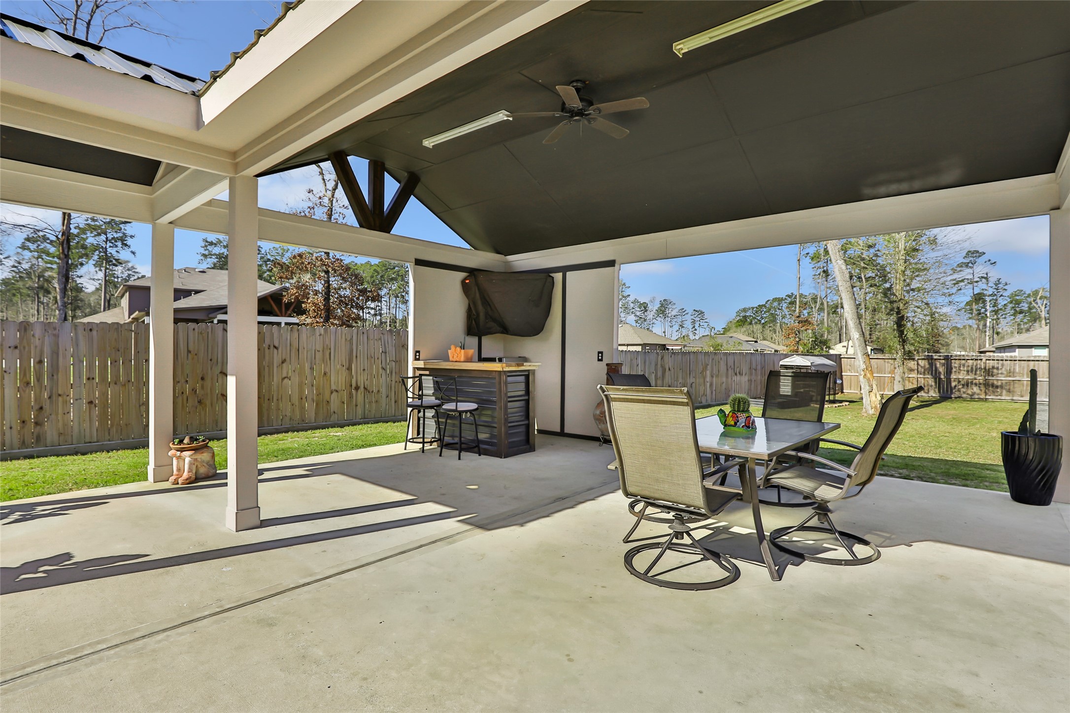 811 Republic Road New Caney, TX 77357 - Photo 7 of 37 This photo showcases a spacious covered patio, perfect for outdoor relaxation and entertainment. It features a ceiling fan, a dining area, and a bar setup, all overlooking a well-maintained yard with privacy fencing.