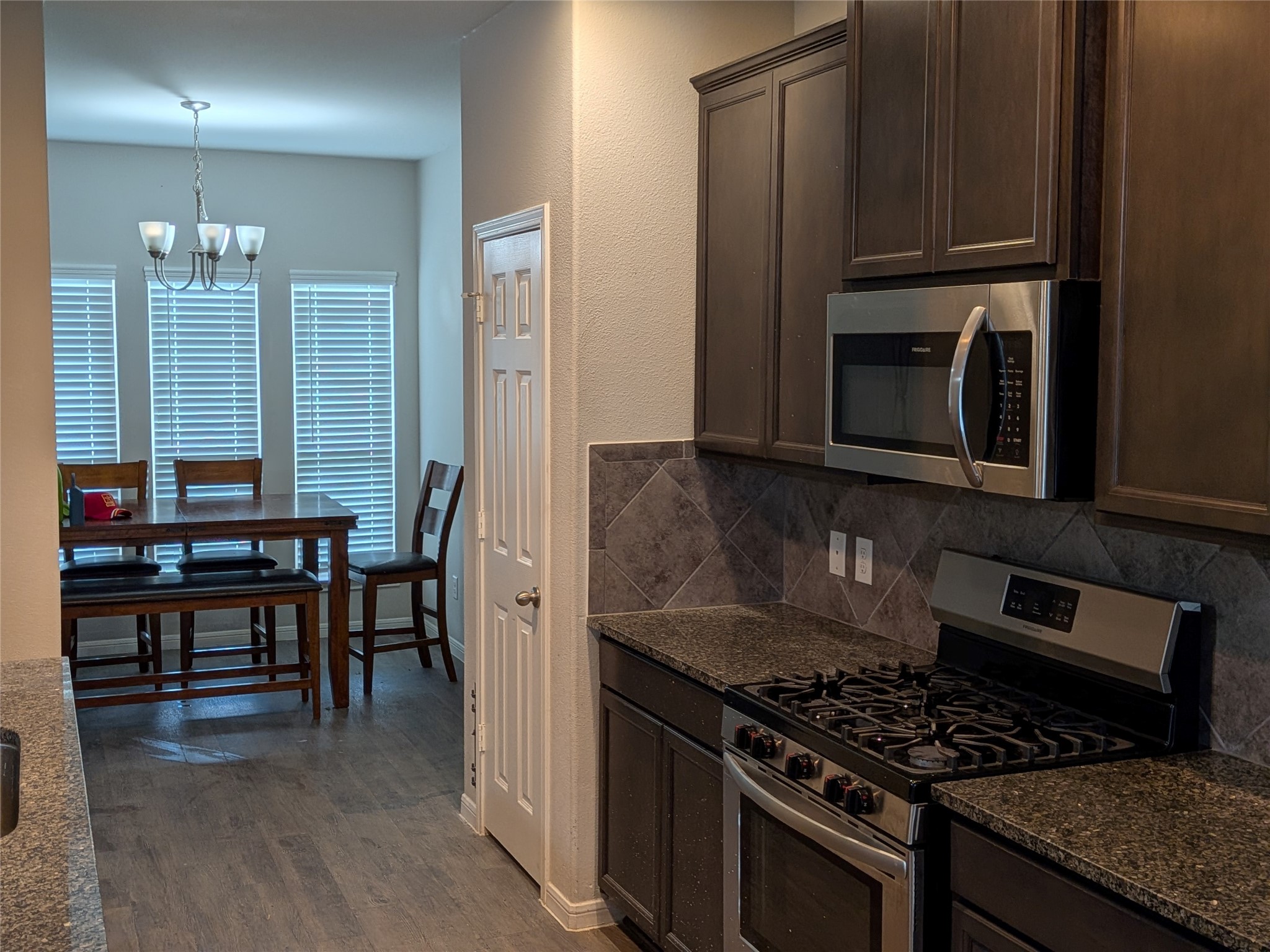 15730 Joe Dimaggio Street Splendora, TX 77372 - Photo 7 of 20 This kitchen features dark cabinetry, granite countertops, and stainless steel appliances, including a gas stove and microwave. A dining area is visible with large windows and a modern chandelier, providing a bright and welcoming space.