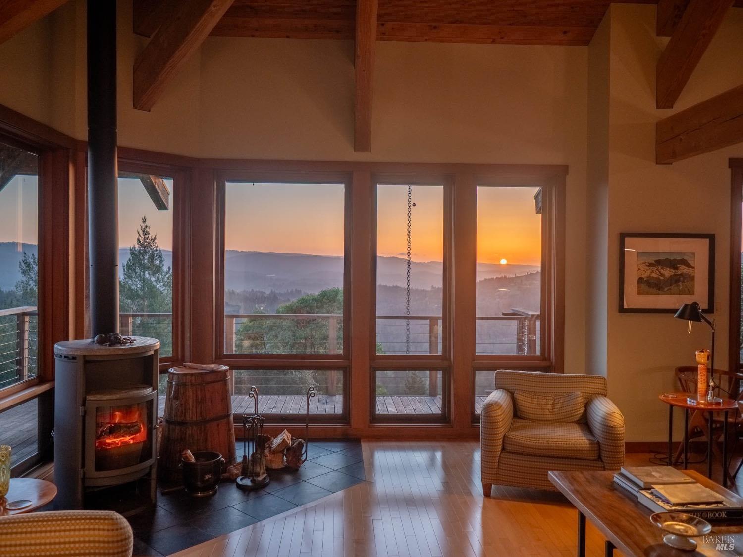 10951 Big Meadow Road Philo, CA 95466 - Photo 5 of 46 Golden-orange hues stretch across the horizon, casting a soft glow on the wooden floors and bathing the entire room in a honeyed light. Through the glass doors and wraparound windows, you can see layers of distant hills fading into dusky blues with the sun resting gently above.