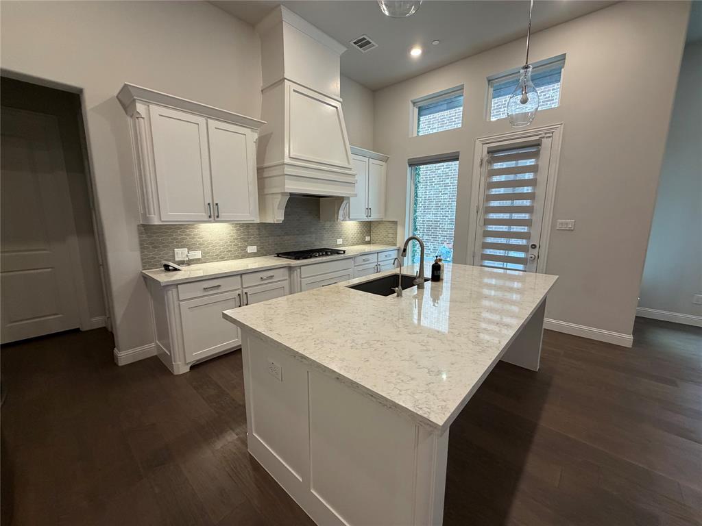 1049 Sarah Street Allen, TX 75013 - Photo 5 of 12 Kitchen featuring a large central island with a built-in sink, white shaker-style cabinetry, light-toned countertops, a decorative range hood, and dark wood-finish flooring
