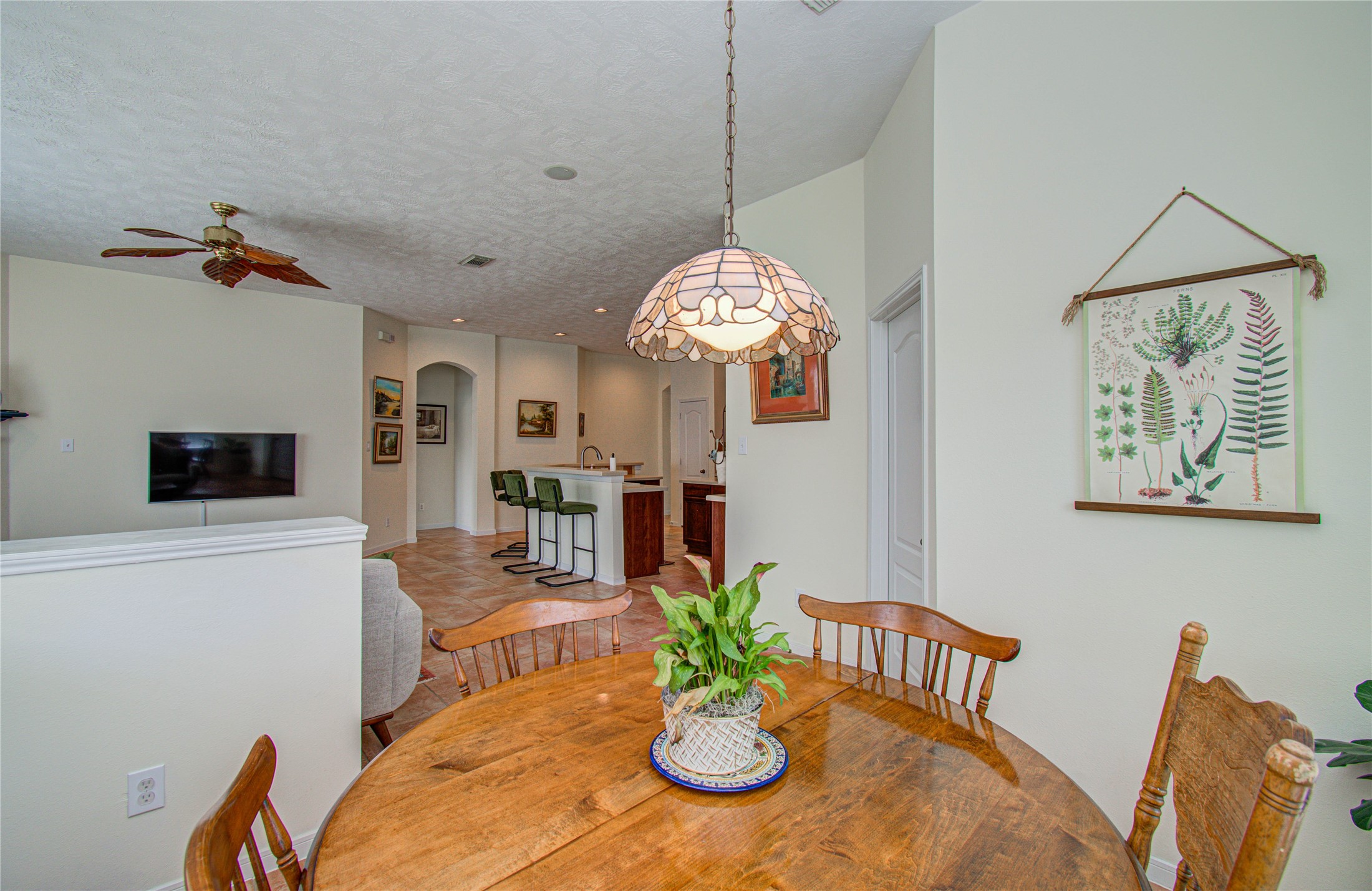 20007 Alcea Court Spring, TX 77379 - Photo 18 of 36 This cozy dining area features a wooden table with chairs, a decorative hanging light, and a wall with botanical art. It opens into a living space with a ceiling fan and a wall-mounted TV, leading to a kitchen with bar seating, creating a warm, inviting atmosphere.
