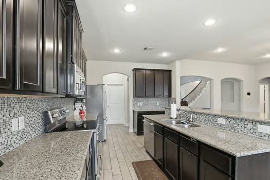 24714 Longwood Forest Drive Spring, TX 77373 - Photo 8 of 21 Modern kitchen with dark cabinets, granite countertops, and stainless steel appliances. Features a mosaic tile backsplash, spacious layout, and recessed lighting, leading to an open living area with archways.