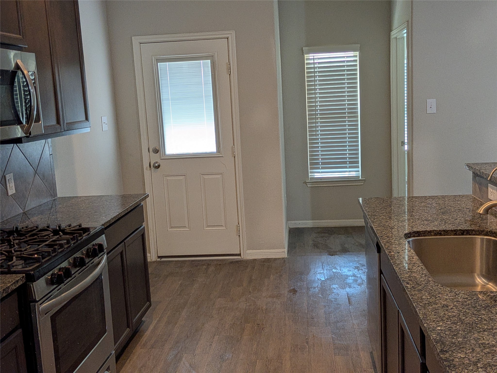 15730 Joe Dimaggio Street Splendora, TX 77372 - Photo 12 of 20 This kitchen features dark wood cabinetry, granite countertops, a stainless steel gas stove, and a microwave. It has a tiled backsplash, a double sink, and a door leading to the outside. Natural light enters through a window with blinds.