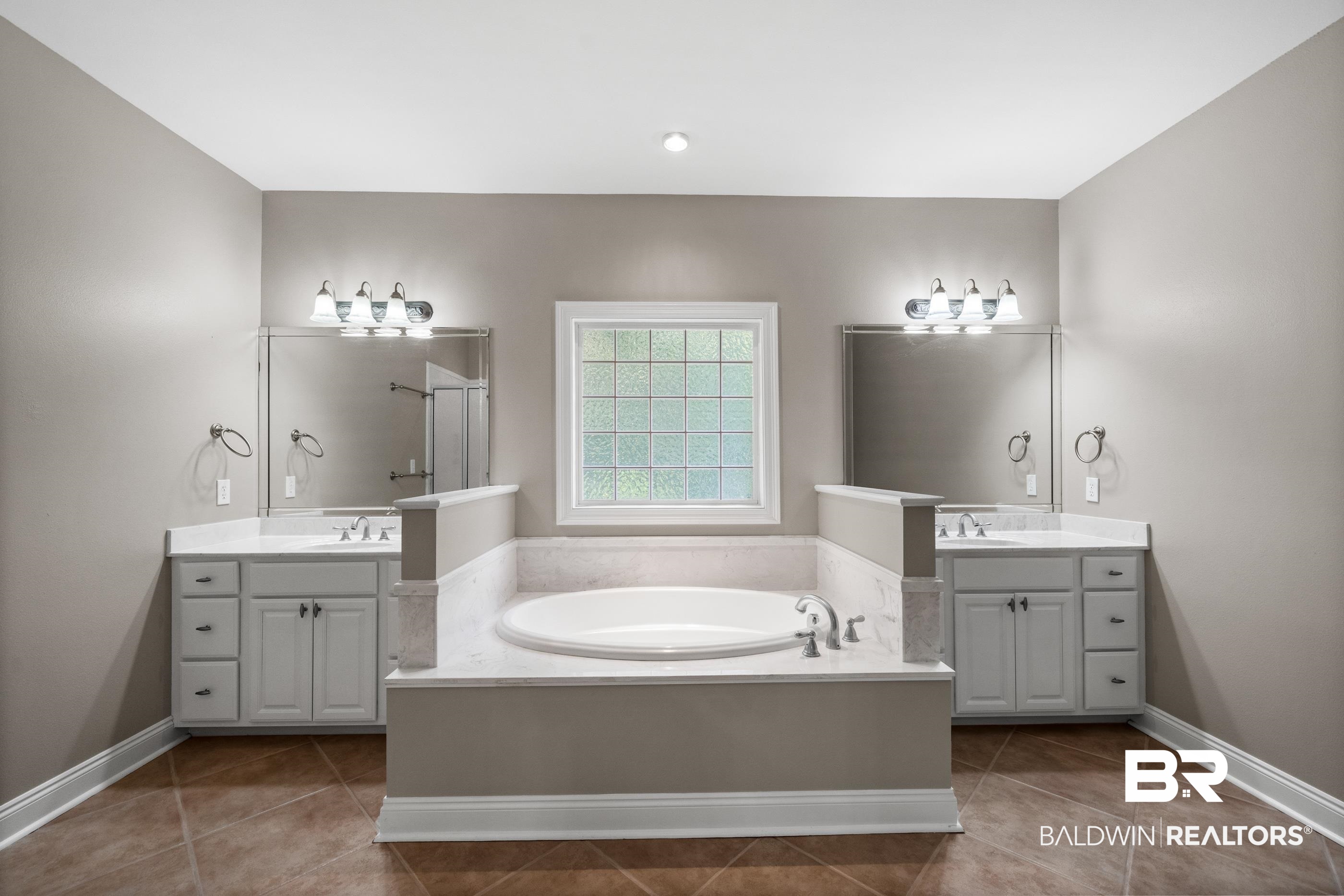 303 Savannah Circle Foley, AL 36535 - Photo 21 of 34 Central soaking tub with surrounding white-finish countertop, flanked by dual vanities with integrated sinks and wall-mounted light fixtures, set on a tile floor