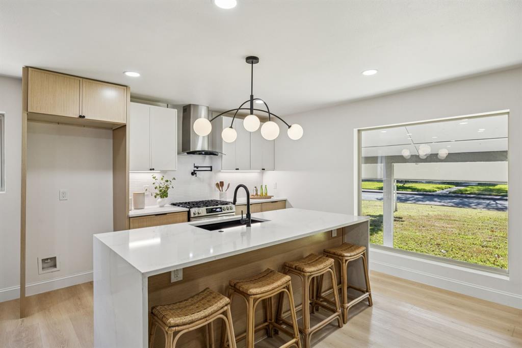 3115 Whirlaway Road Dallas, TX 75229 - Photo 6 of 28 Kitchen featuring light wood-style flooring, a kitchen bar, decorative backsplash, wall chimney range hood, and light stone countertops
