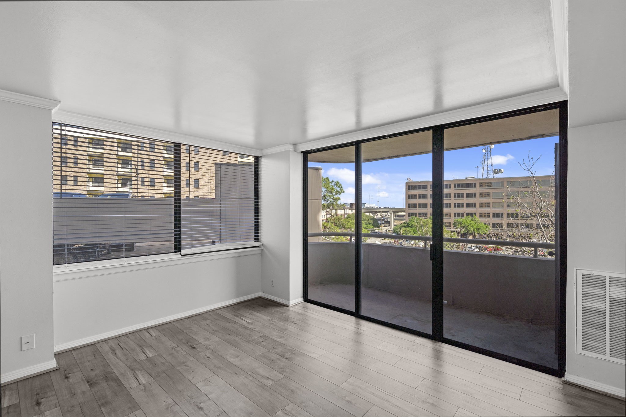 A bright living room with large windows and sliding glass doors leading to a balcony. The space features vinyl flooring, and ample natural light with urban views.