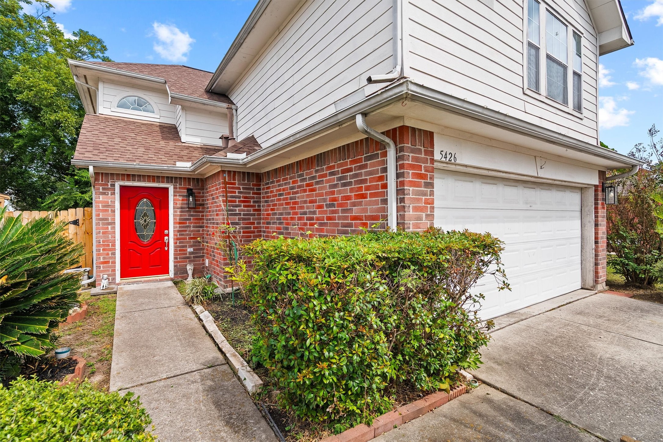 Captivating two-story home with a striking red front door, classic brick facade, and spacious attached garage, nestled in a lush, landscaped setting.