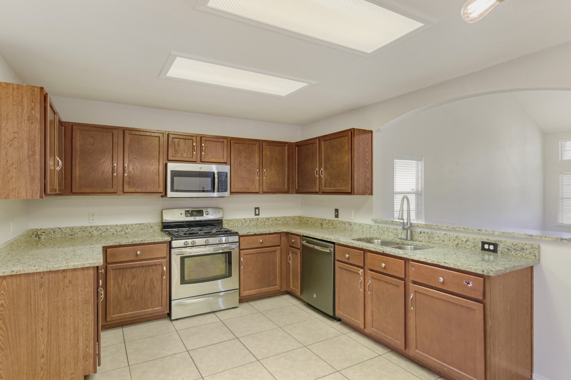 4010 Reeders Drive Austin, TX 78725 - Photo 14 of 26 Kitchen featuring appliances with stainless steel finishes, light stone countertops, brown cabinetry, light tile patterned flooring, and a peninsula