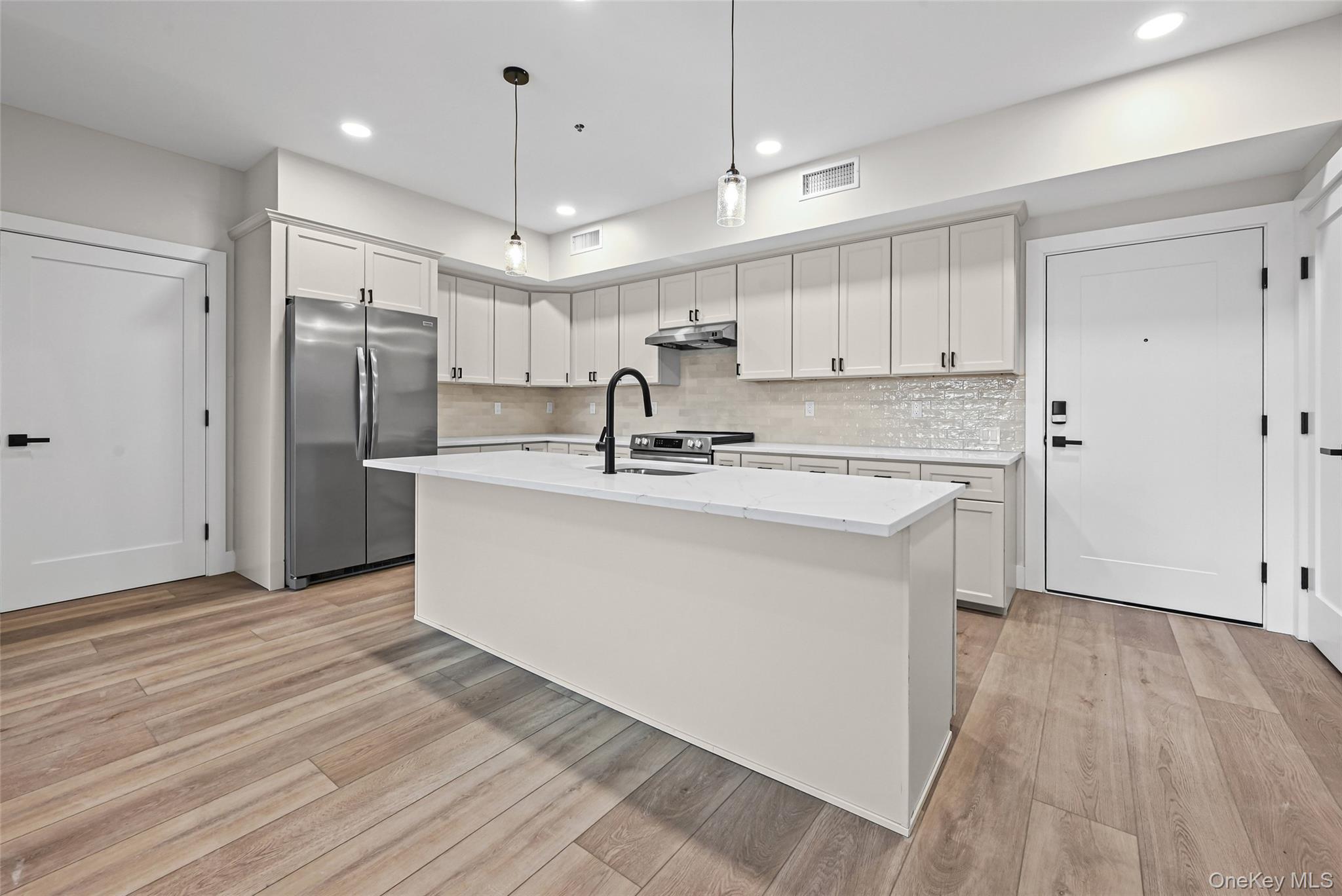 80 Division Street, Unit 3A Patchogue, NY 11772 - Photo 6 of 18 Kitchen featuring appliances with stainless steel finishes, a center island with sink, white cabinetry, pendant lighting, and light wood-style floors
